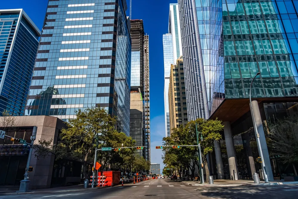 Colorado Street canyon in downtown Texas city, glass tower facades reflecting sky, a building under active construction rising between them