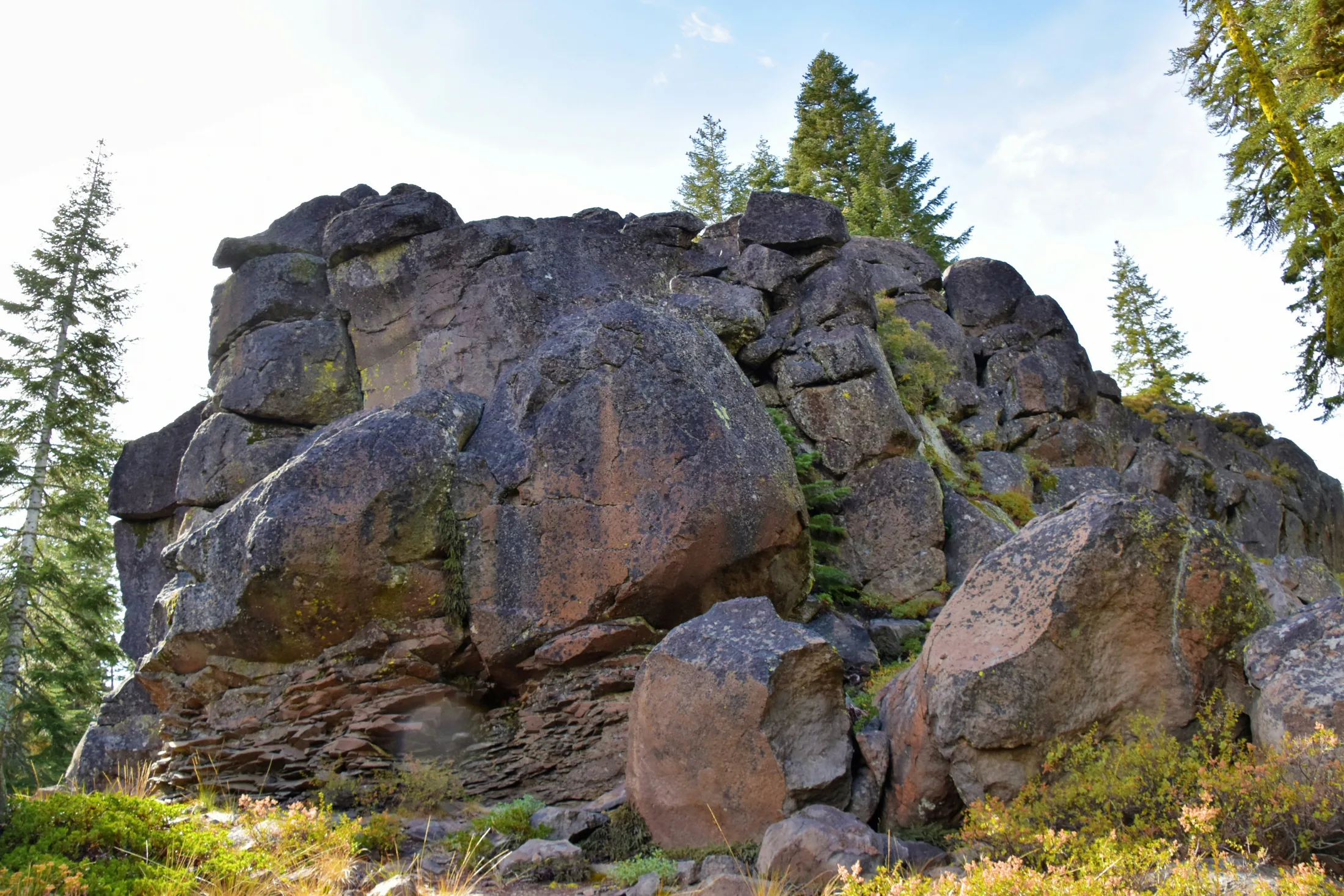A large mass of dark volcanic rock boulders rises above scrubby vegetation with conifers at the ridgeline