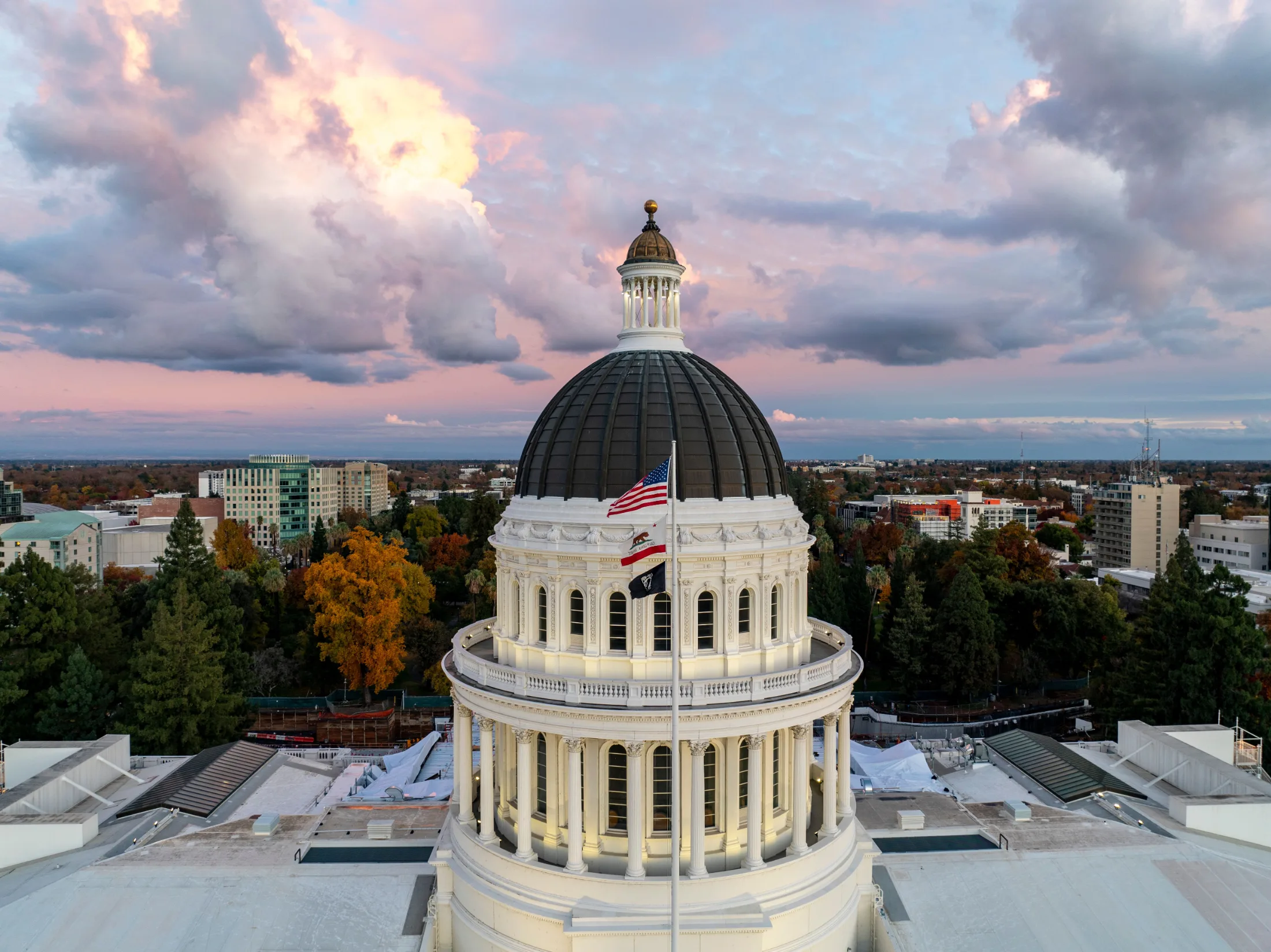 California State Capitol dome at dusk, American and California flags flying, Sacramento skyline in the background