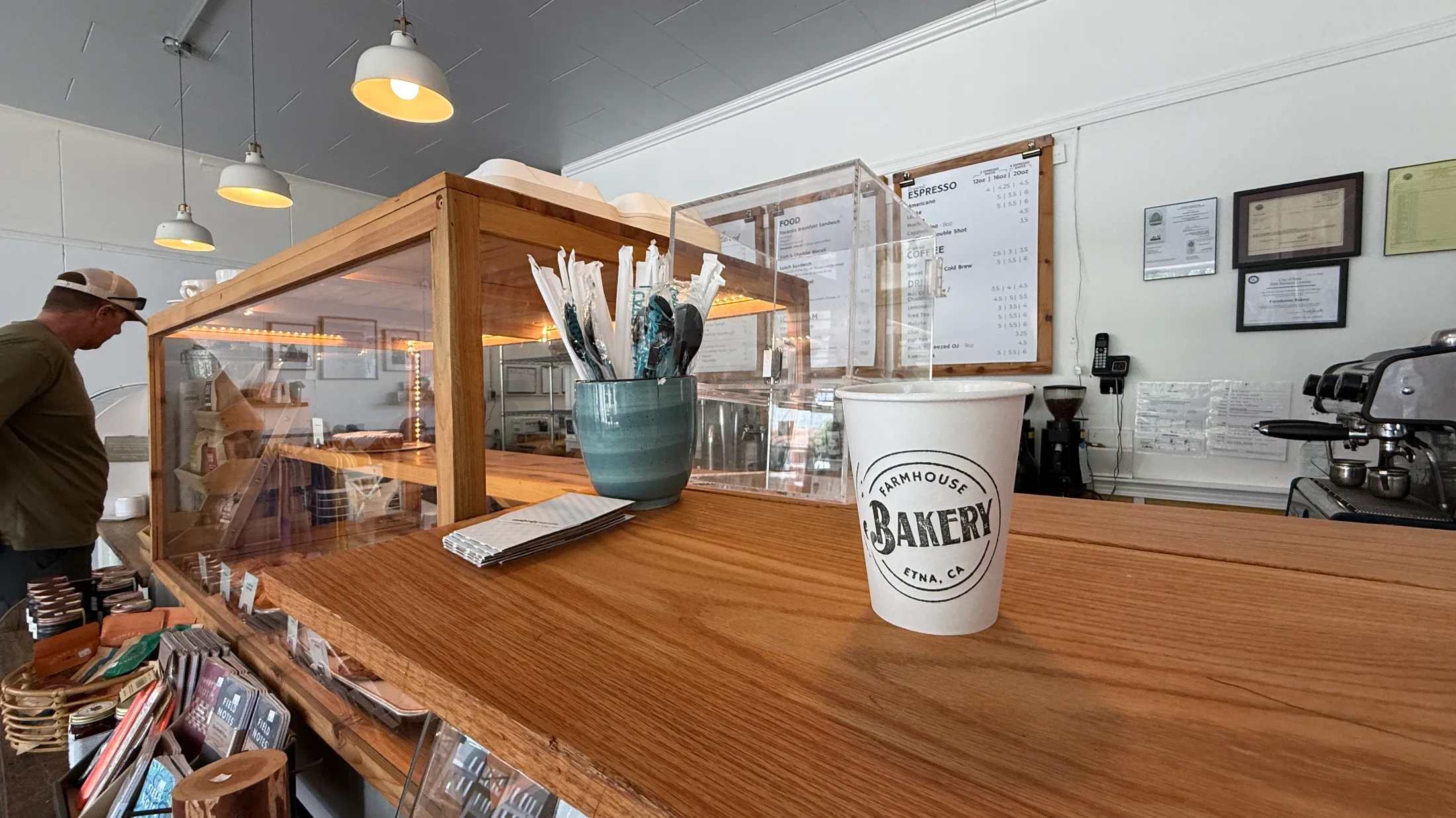 Farmhouse Bakery branded cup on the wooden counter with the espresso machine and menu board visible in the background