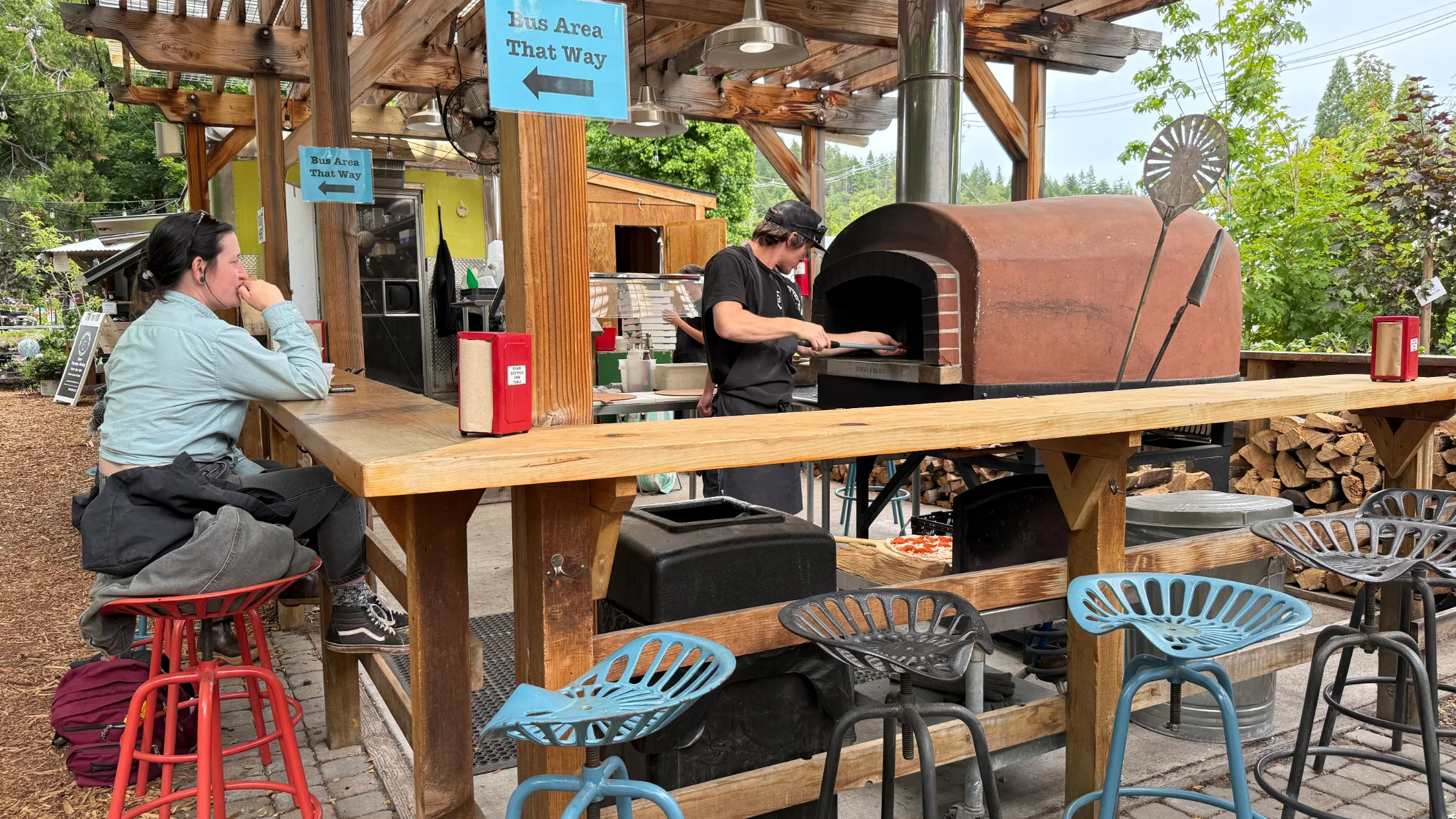Cook working a wood-fired brick oven at the Garden Tap pizza station, with a guest seated at the bar counter
