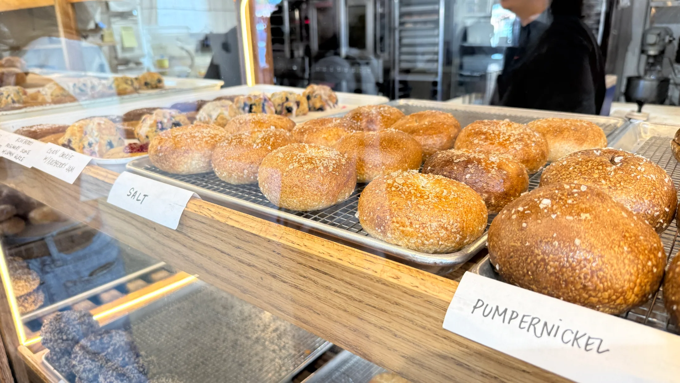 Display case at Sunday Bagels showing trays of salt and pumpernickel bagels with handwritten labels, blueberry pastries visible in the background