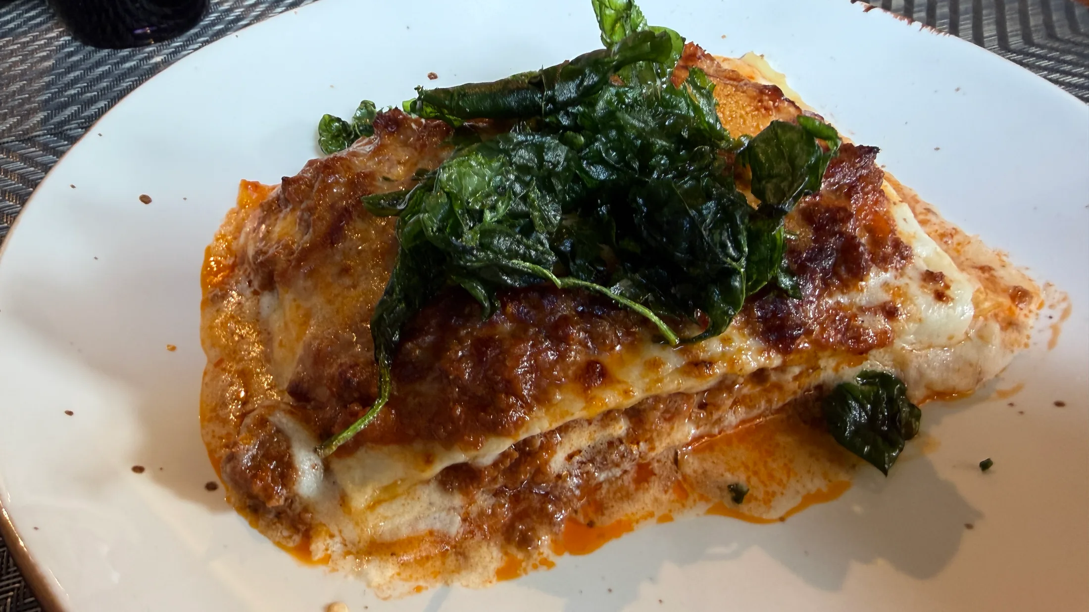 A wedge of lasagna di carne topped with fried spinach, caramelized and blistered edges visible through layers of beef ragù and béchamel