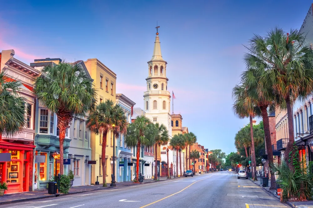 Charleston street at golden hour — colorful historic facades, palm trees, and a white steeple rising against a soft pink-blue sky