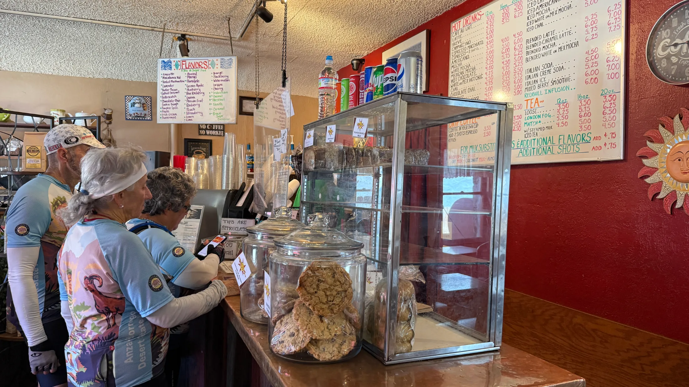 Cyclists in riding gear ordering at the Seven Suns counter, with large cookie jars and the full drink menu board visible