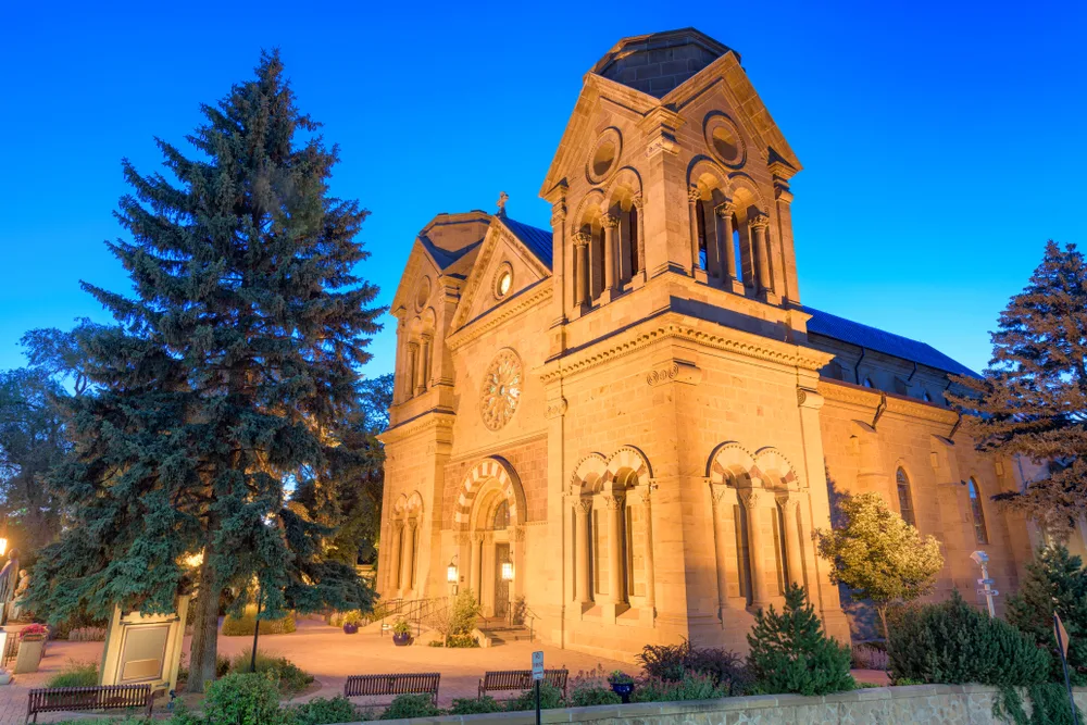 Cathedral Basilica of St. Francis of Assisi lit warm gold at blue hour, tall evergreens silhouetted, empty benches in foreground