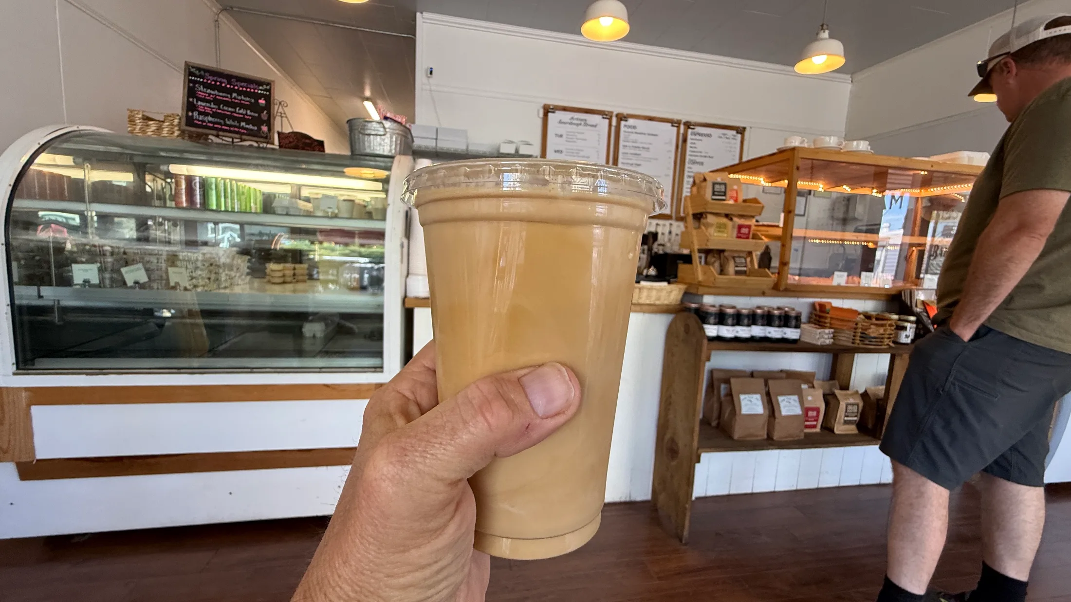 Hand holding a Sweet Cream Cold Brew from Farmhouse Bakery inside the shop, pastry case and espresso menu visible in background