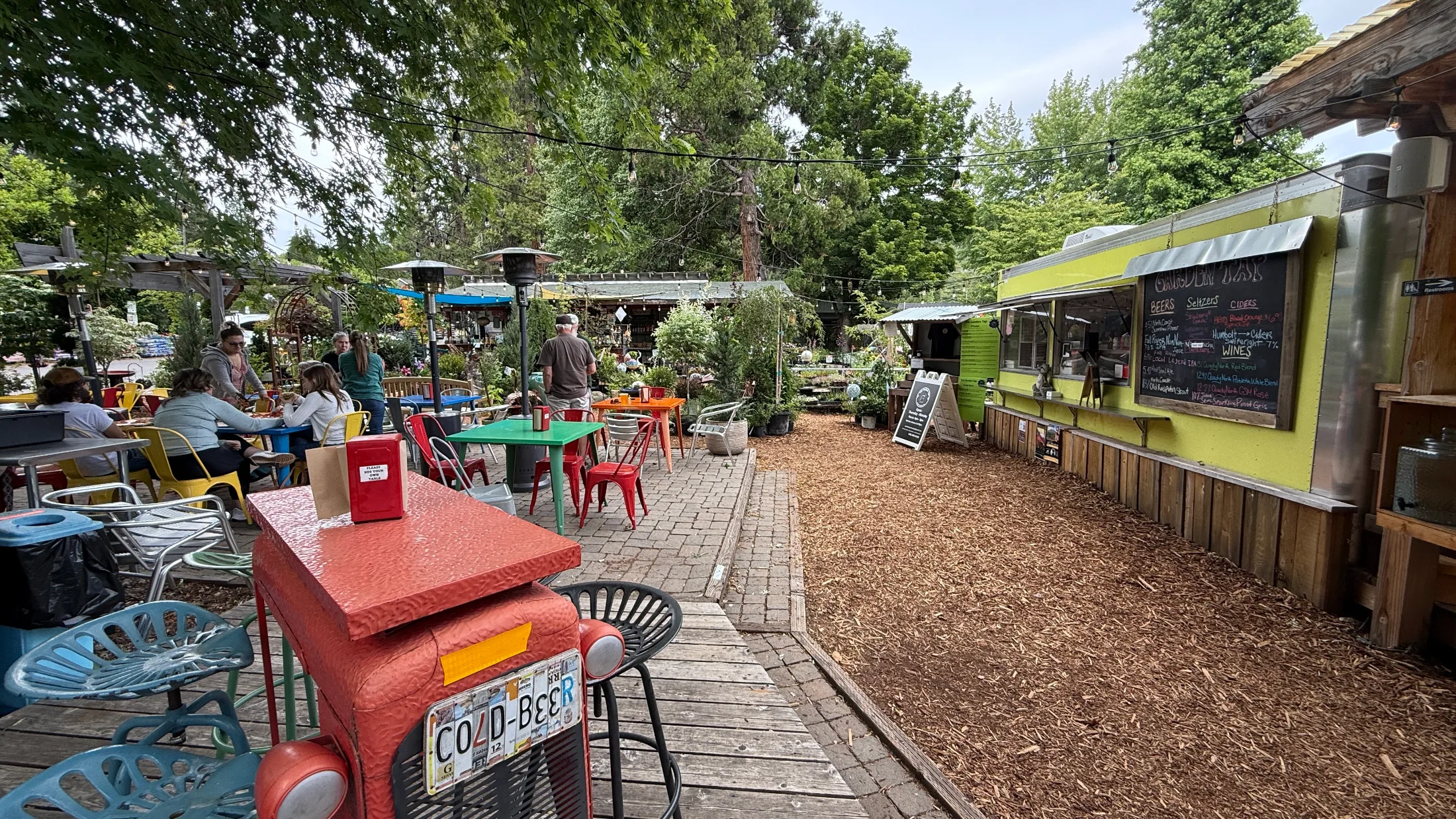 Colorful mismatched tables and chairs fill the outdoor patio at Garden Tap, with a bright green food truck and chalkboard menu visible on the right