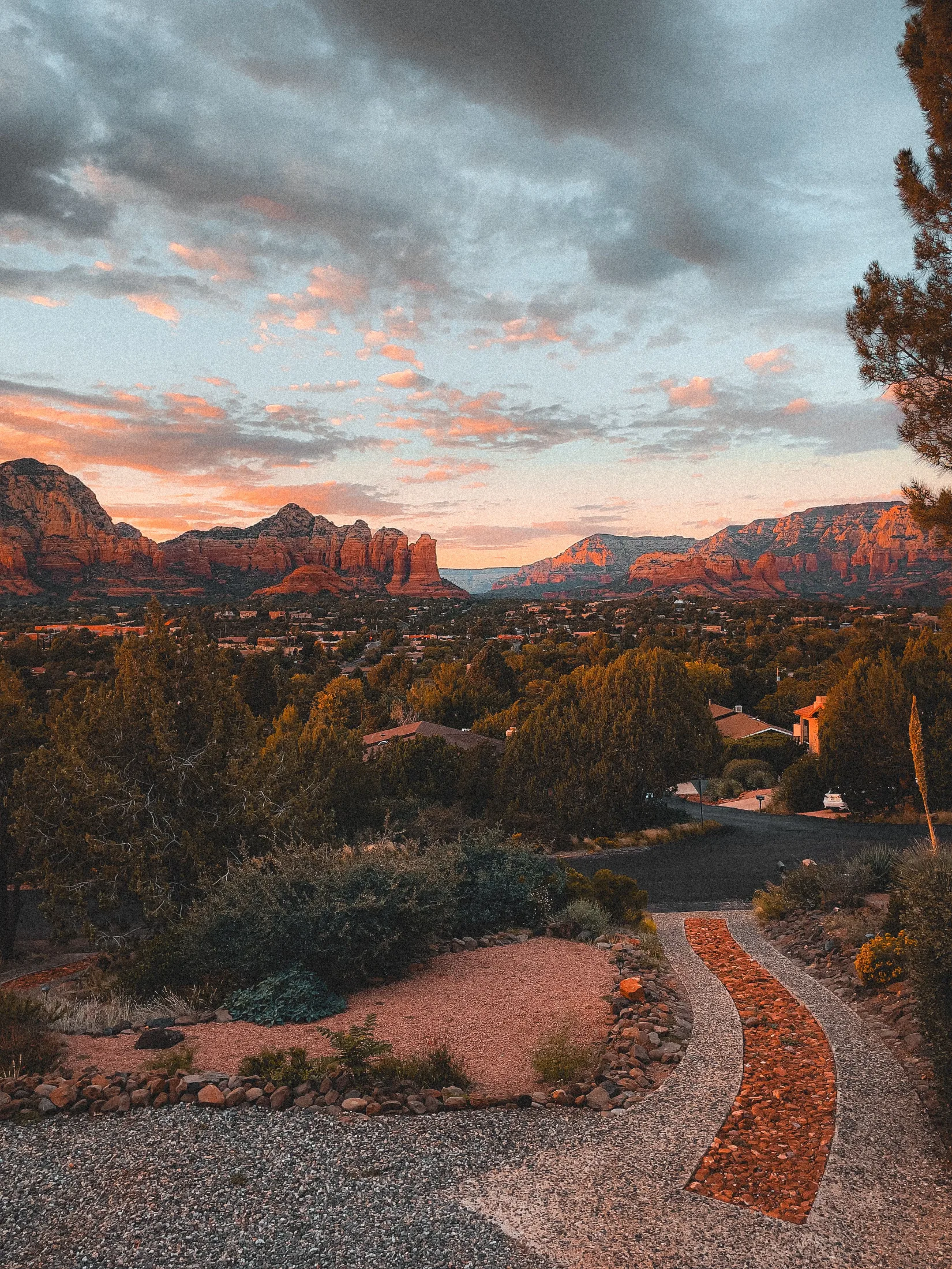 A winding gravel and stone path curves through a desert-landscaped yard at sunset, with Cathedral Rock glowing pink-orange in the distance