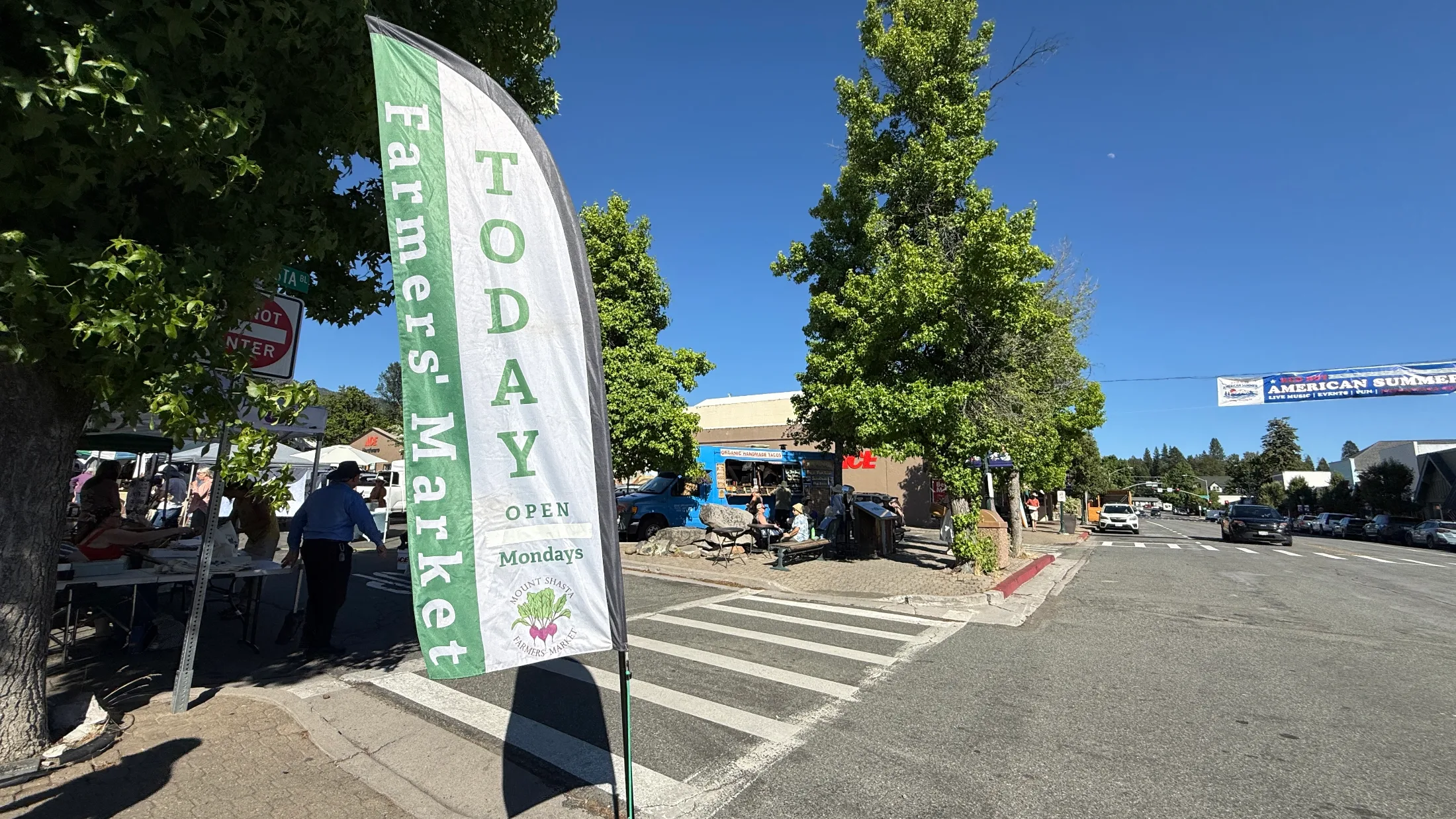 A green and white Farmers Market Today banner flag waves on the Boulevard in downtown Mount Shasta, with the blue organic taco truck and market tents visible behind it.