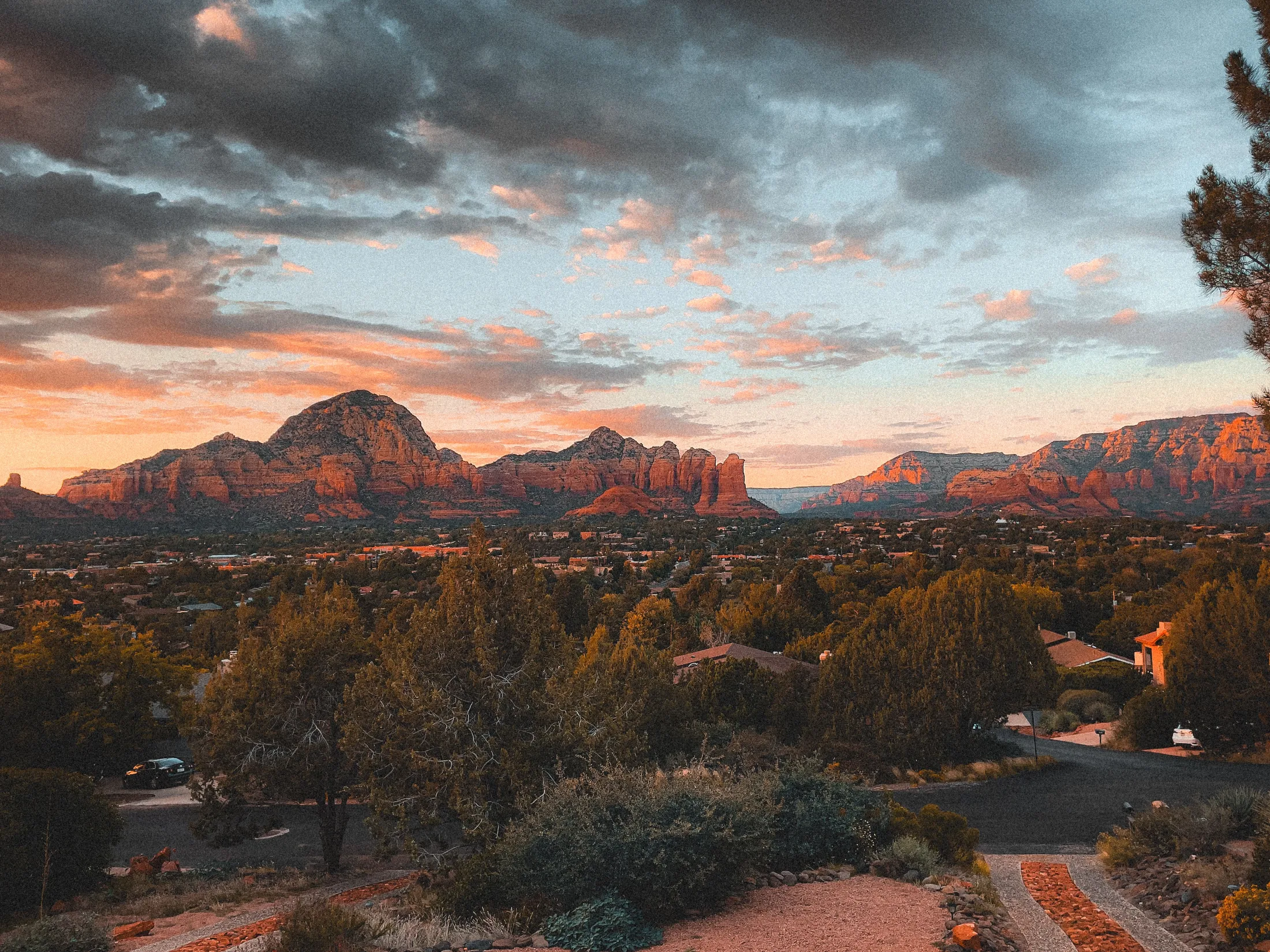 Wide panoramic sunset view across Sedona rooftops and juniper canopy toward glowing red buttes beneath a dramatic cloud-streaked sky