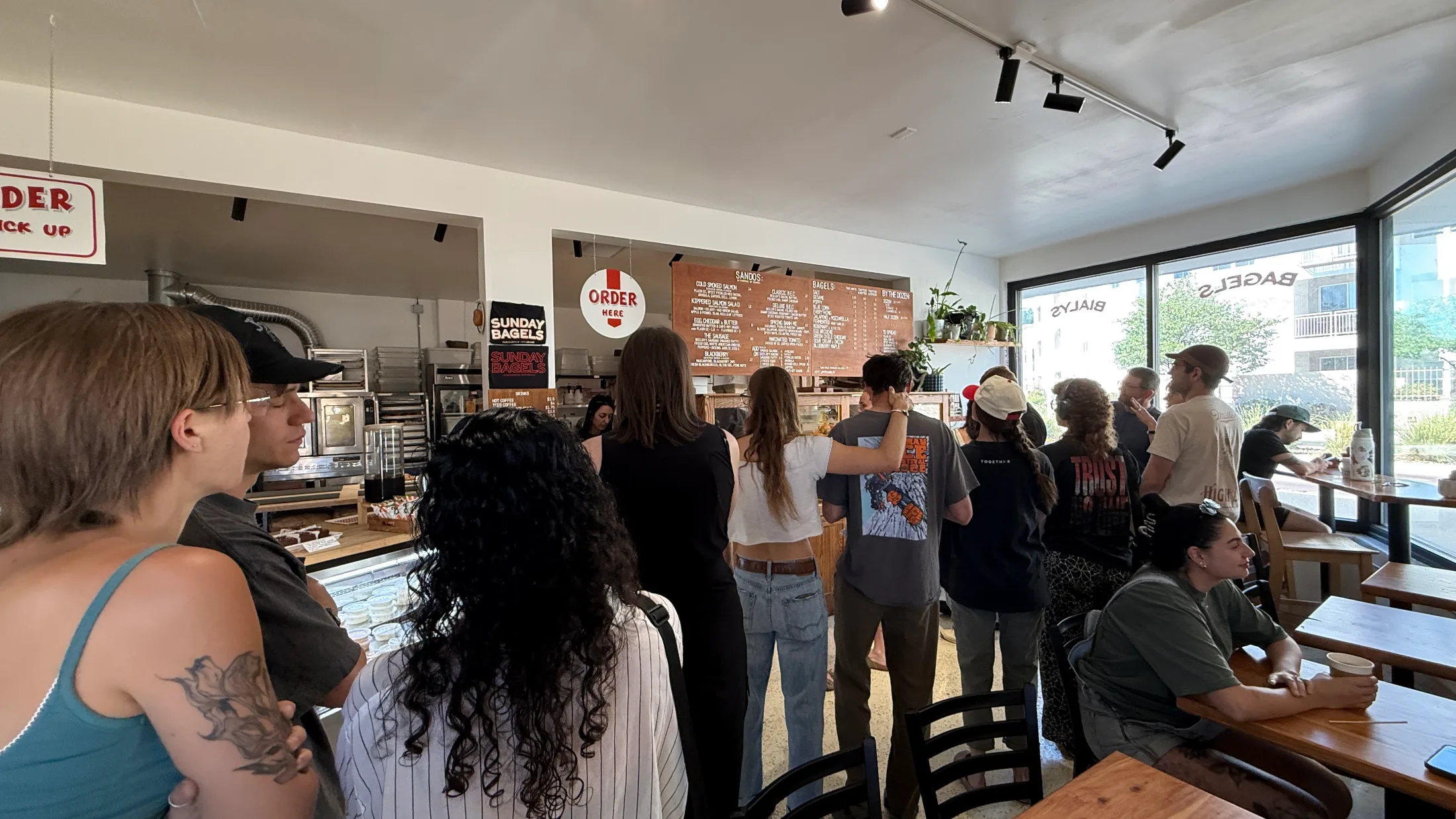 Packed interior of Sunday Bagels in Albuquerque, customers lined up at the order counter with the full menu board visible on the wall