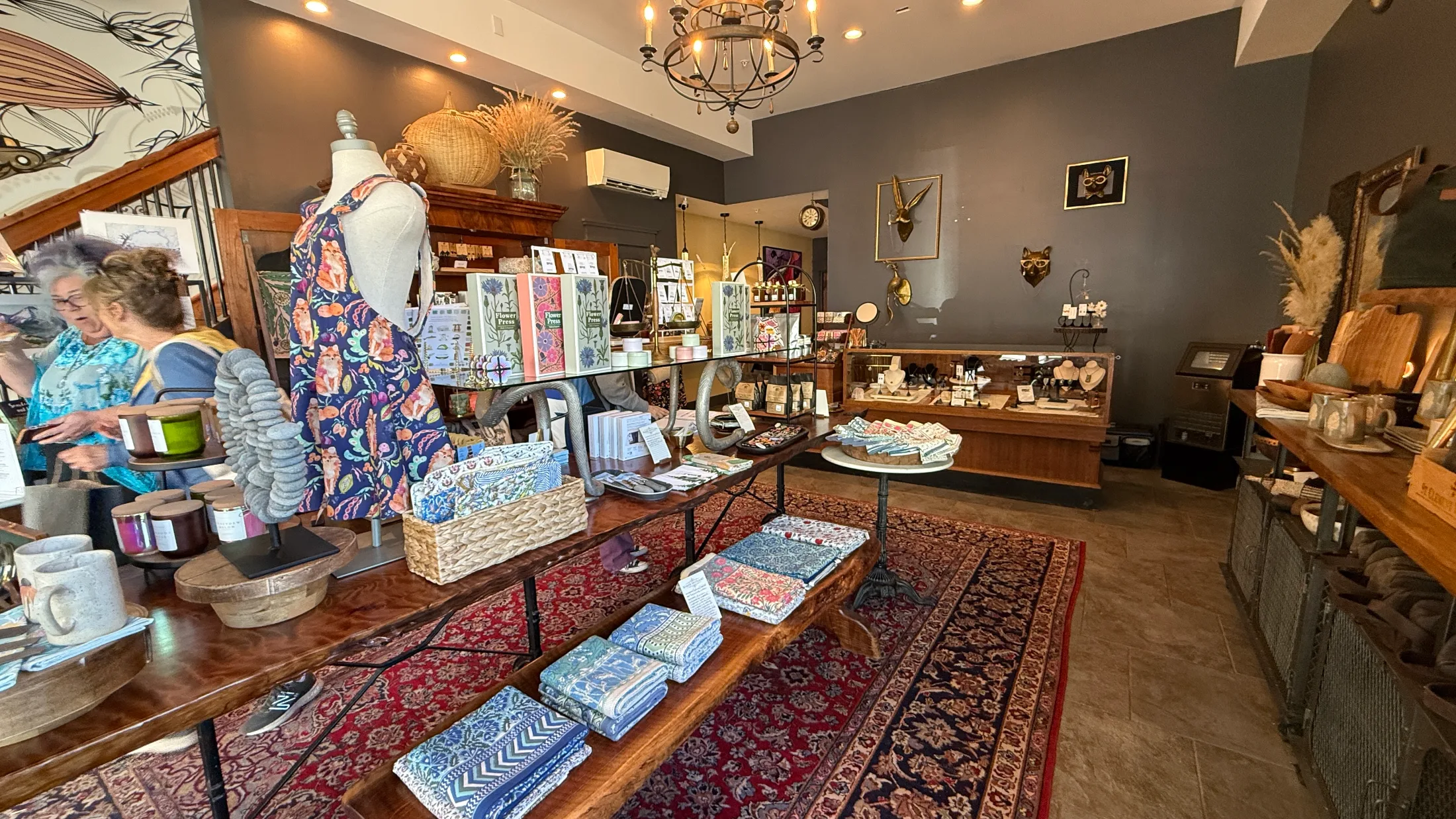 Interior of the Mossbrae Keepshop showing curated goods on walnut tables over a red Persian rug, with guests browsing