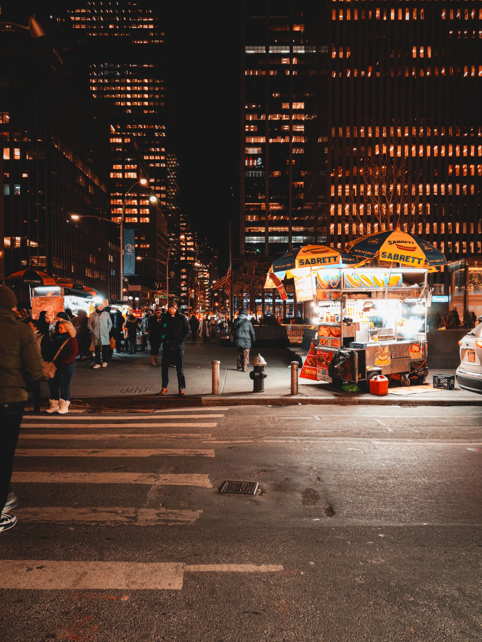 A row of Sabrett hot dog and pretzel carts lit up at night on a Manhattan avenue, skyscrapers glowing orange in the background, pedestrians gathered on the corner