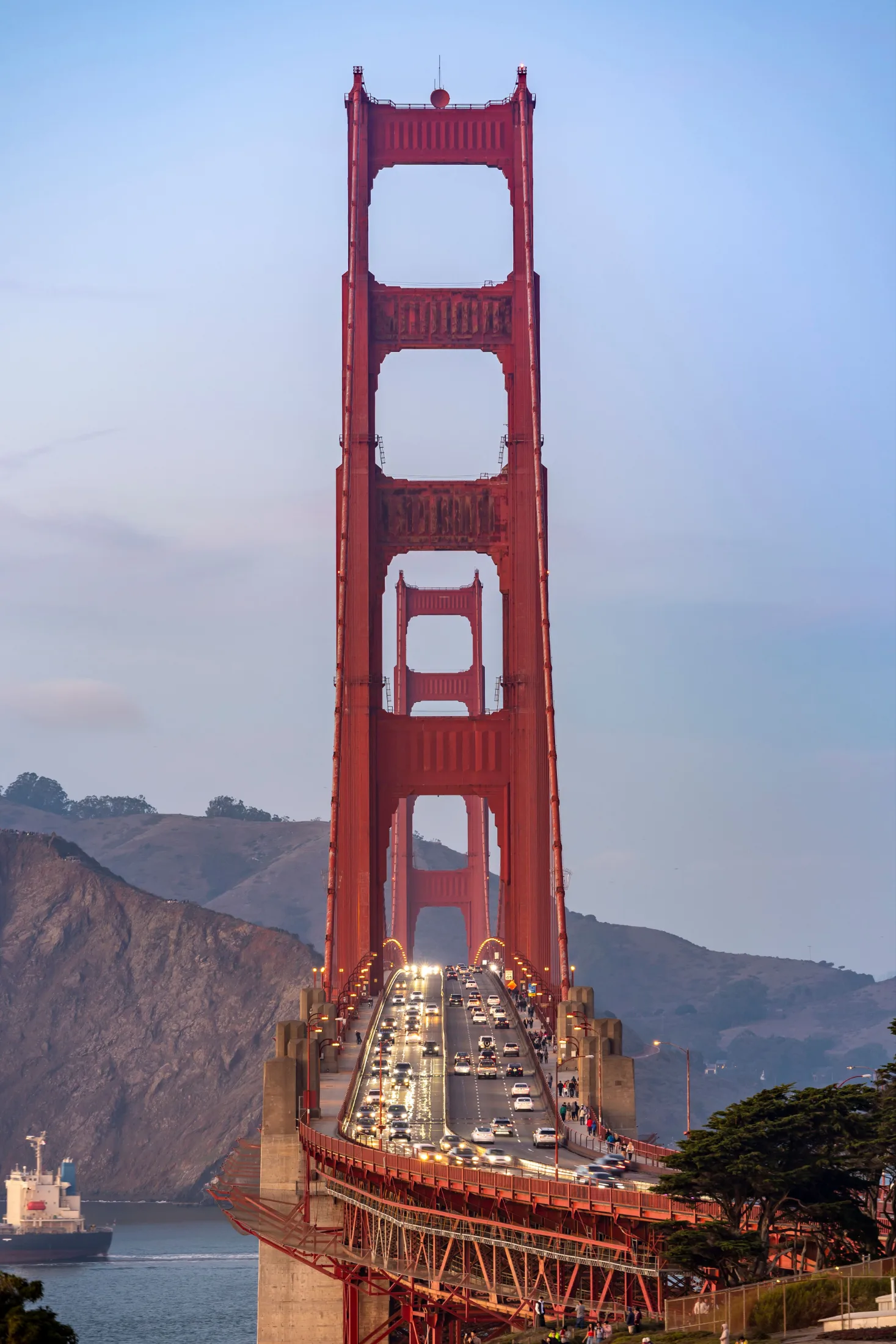 Golden Gate Bridge tower at dusk with headlights streaming across the span and the Marin Headlands behind