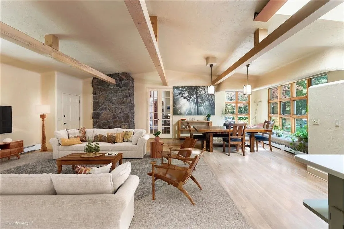 Interior living and dining area with exposed wood beams, stone fireplace, hardwood floors, and large grid windows
