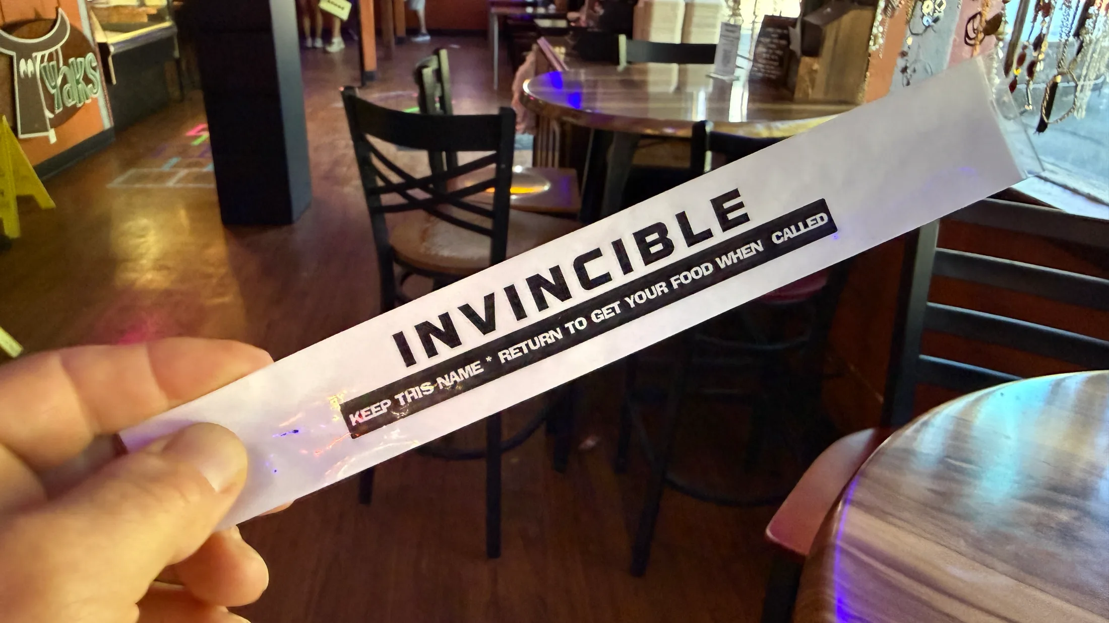 A hand holding a translucent order name tag printed with INVINCIBLE inside Yaks on the 5