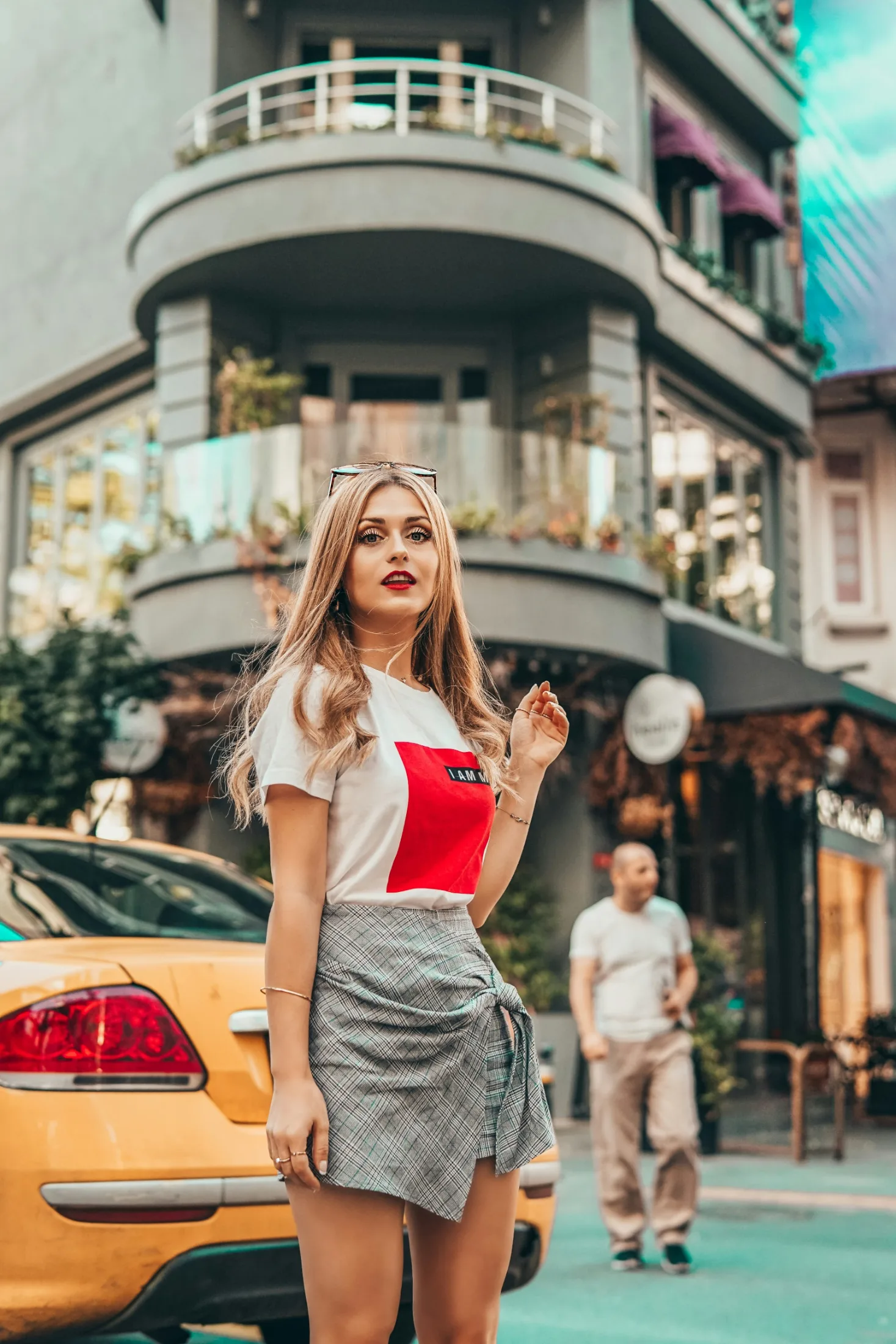 A confident young woman in a graphic tee and plaid wrap skirt stands on a stylish urban block beside a yellow cab