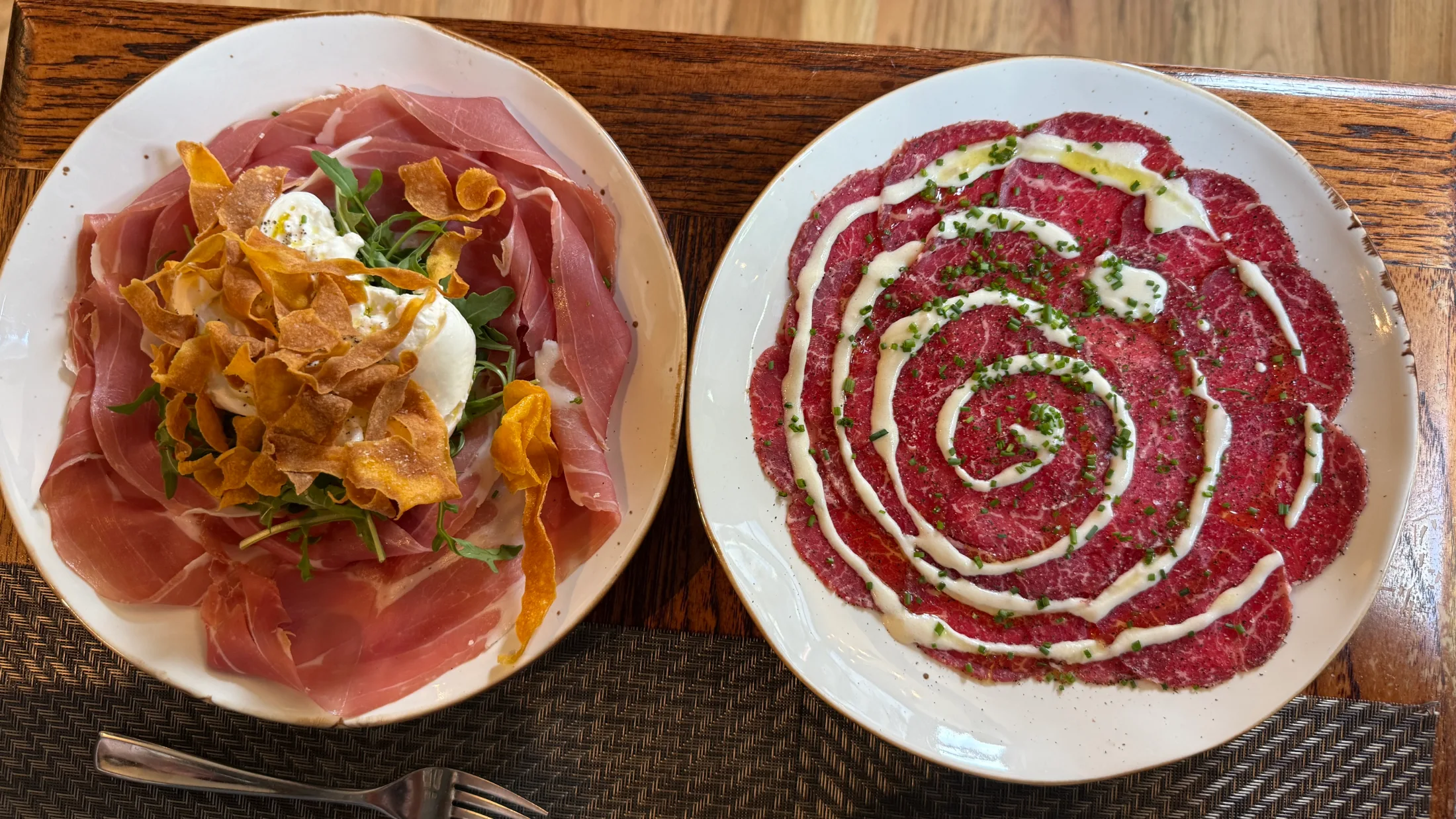 Two antipasti plates side by side: burrata with Parma prosciutto, arugula, and fried pumpkin on the left; carpaccio di manzo with truffle fondue spiraled in concentric circles on the right