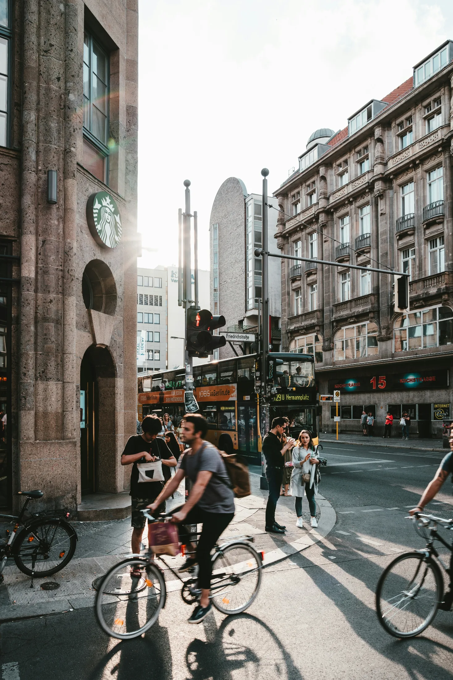 A sunlit European city street at Friedrichstraße filled with cyclists, a double-decker bus, pedestrians, and stone buildings glowing in afternoon light