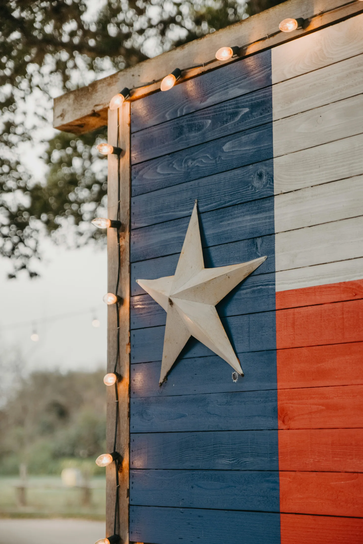 Wooden Texas flag painted in red, white, and blue with a metal Lone Star at center, strung with warm Edison bulb lights at dusk
