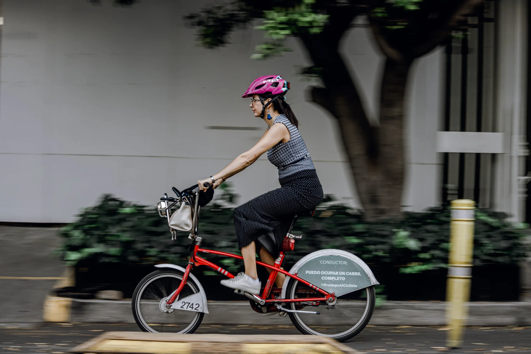 A woman in a skirt and pink helmet rides a red bike-share bicycle in motion through an urban street