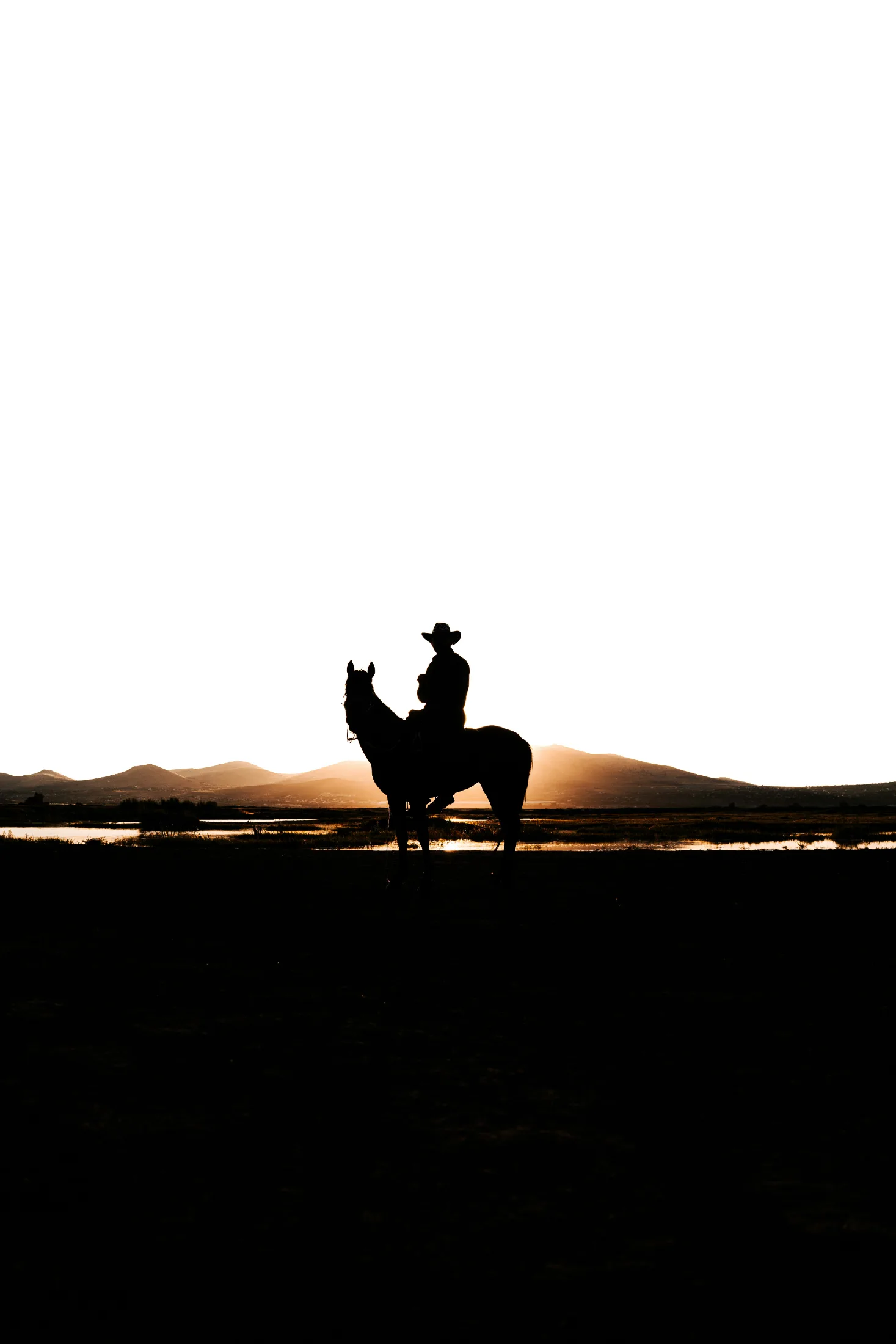 Silhouette of a cowboy on horseback at sunset, mountains and a flat reflective plain behind them