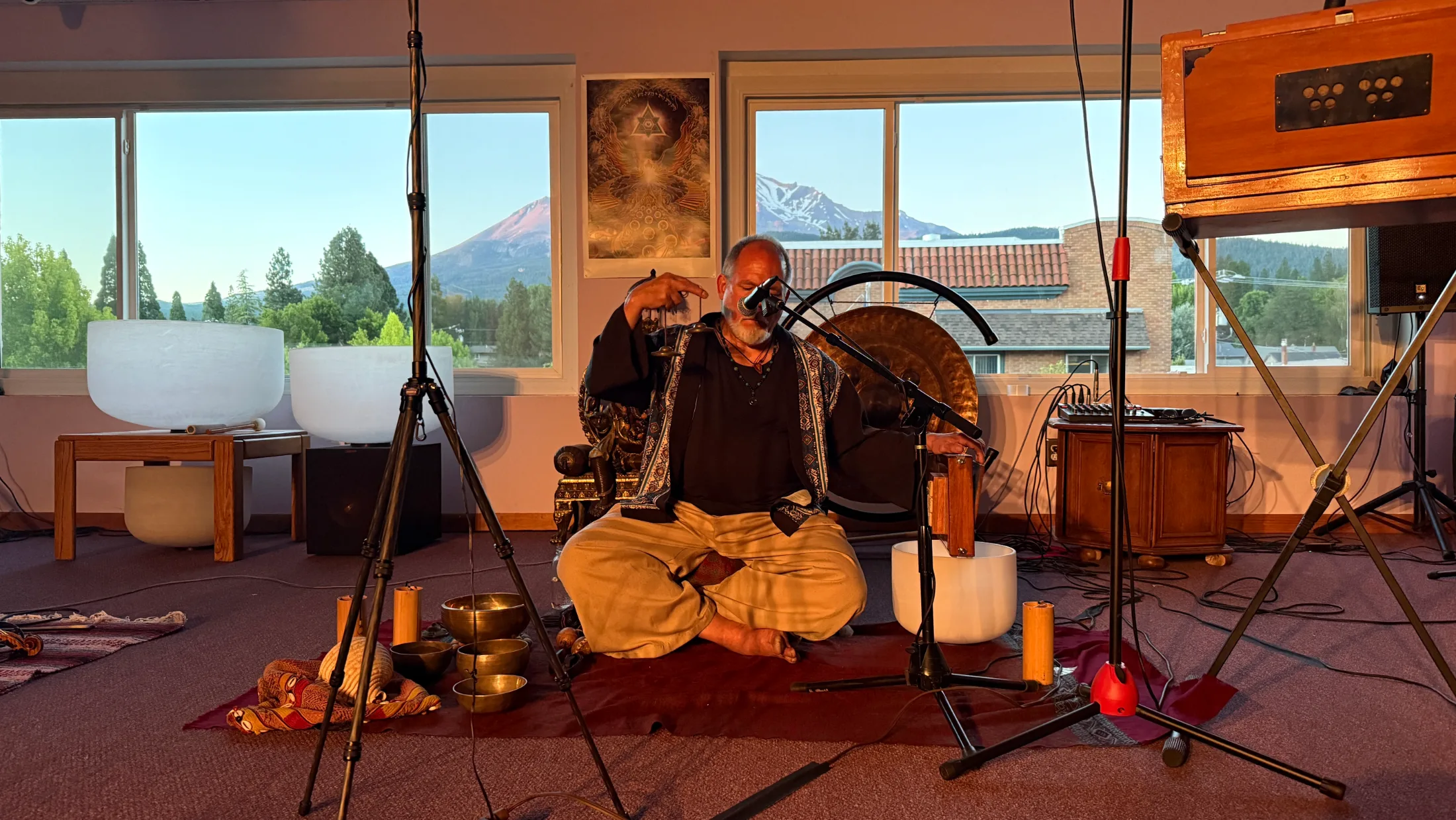 Matthew Kocel seated cross-legged on a red cloth, singing into a microphone surrounded by Tibetan singing bowls and crystal bowls, Mount Shasta glowing orange-pink through the windows at dusk