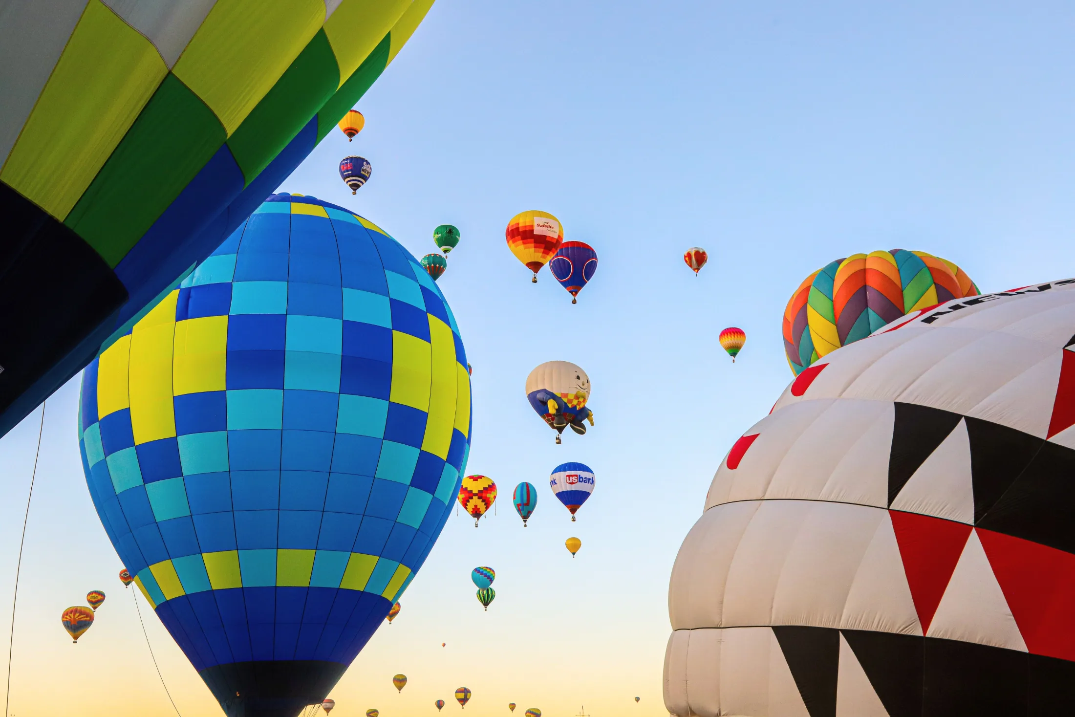 Dozens of colorful hot-air balloons rising into a pale blue morning sky at Balloon Fiesta Park