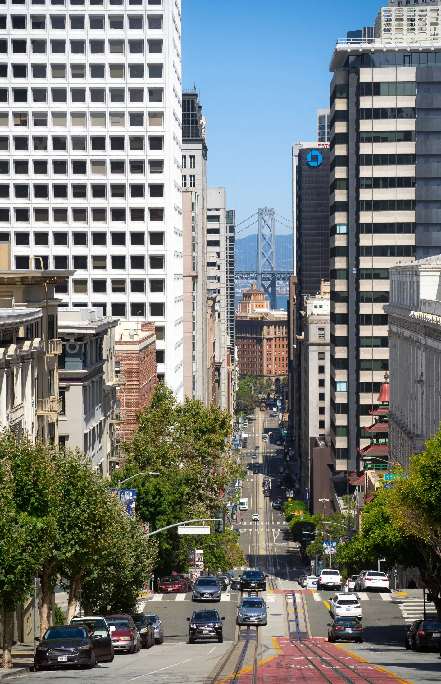 Looking down a steep San Francisco street lined with trees and high-rise buildings, the Bay Bridge visible in the distance
