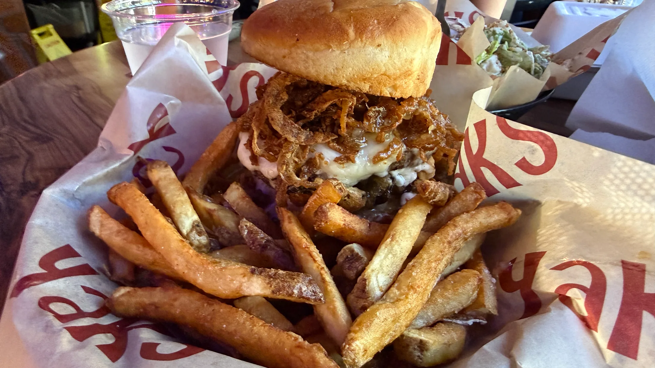 A loaded Yaks burger piled with crispy onion strings and melted cheese, surrounded by hand-cut fries in a branded paper basket