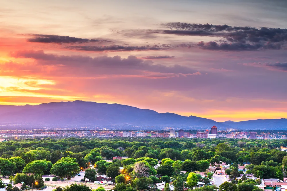 Albuquerque at sunset, tree-lined neighborhoods and downtown towers glowing under a sky of orange and violet, the Sandias purple in the distance