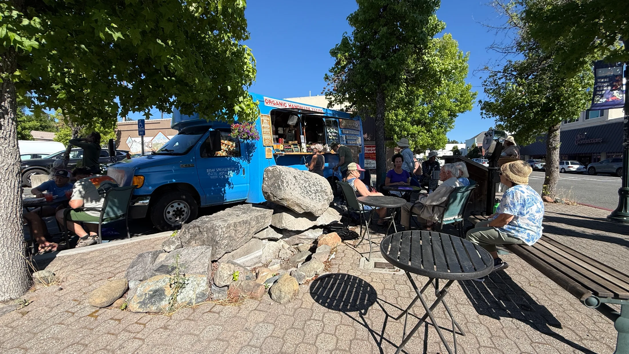A bright blue organic handmade taco truck parked on the cobblestone median of the Boulevard, surrounded by plane trees, with people eating at cafΓ© tables nearby.