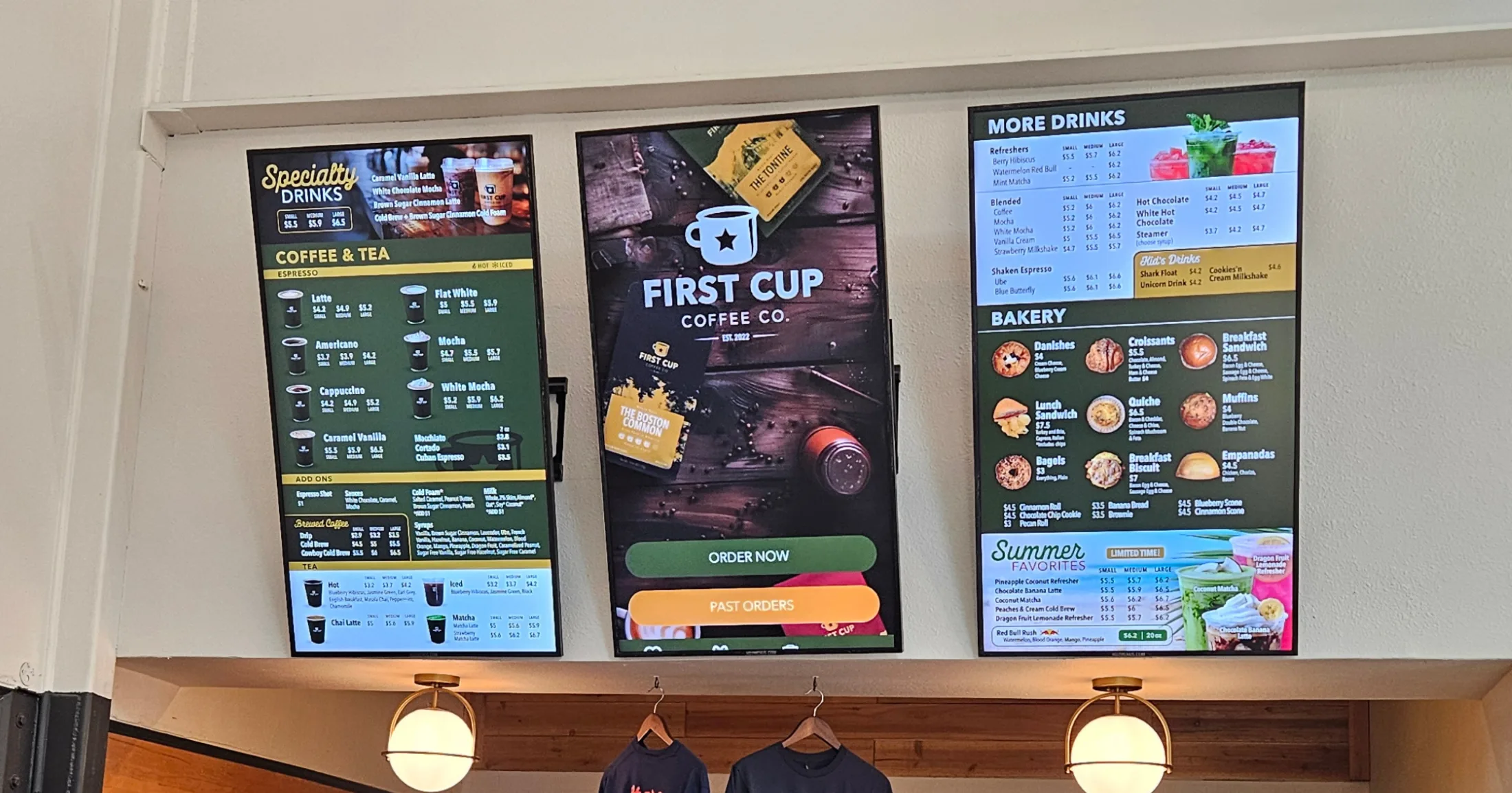 Digital menu boards at First Cup Coffee Co. showing specialty drinks, coffee and tea selections, bakery items, and pricing