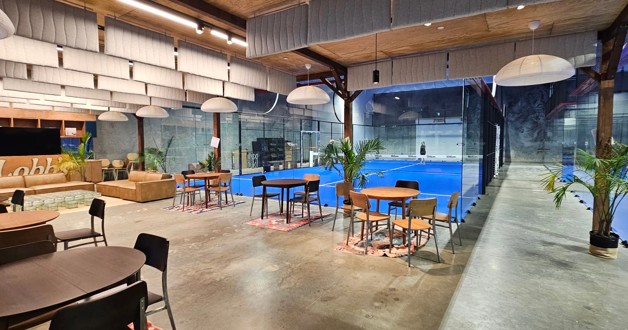 Lobb's Padel lounge and café area with round café tables, leather sofas, layered rugs on polished concrete floors, and palm plants, looking out onto the blue courts through floor-to-ceiling glass