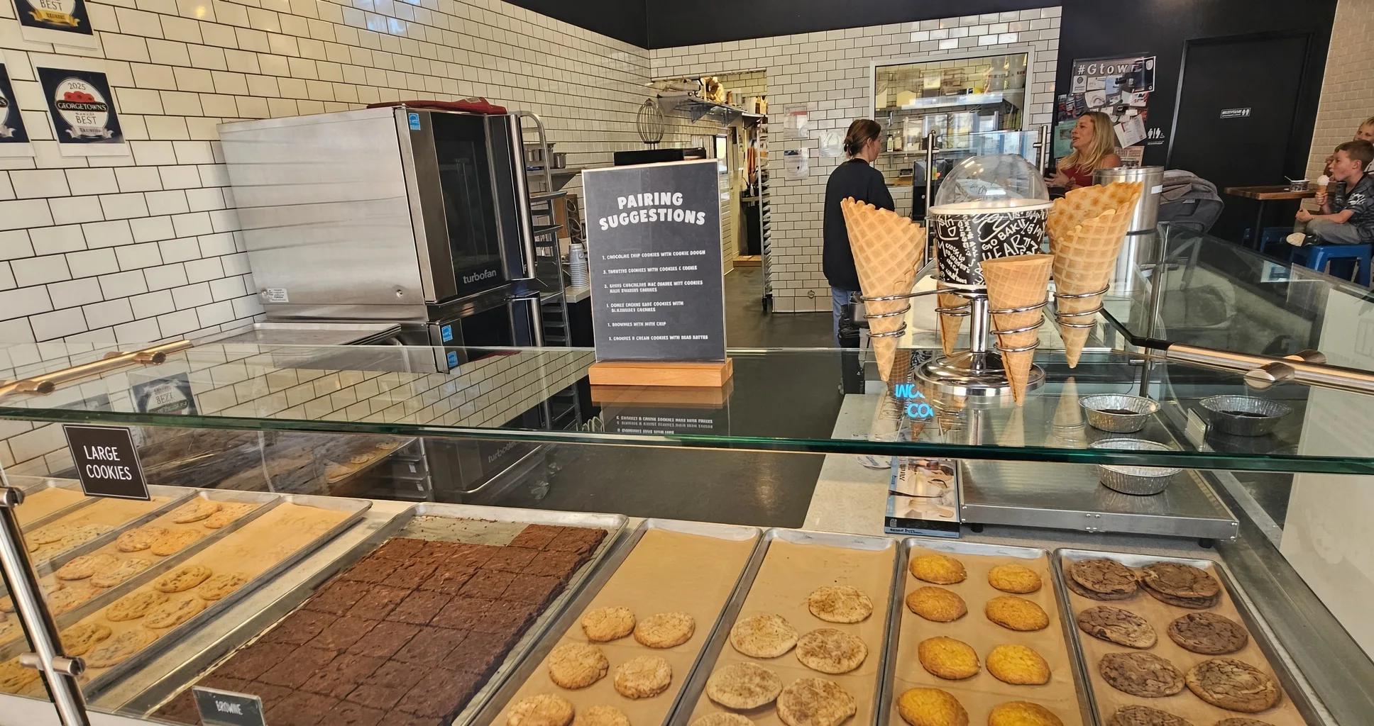 Counter view at The Baked Bear showing large cookies, brownies, snickerdoodles, and a pairing suggestions board near waffle cones