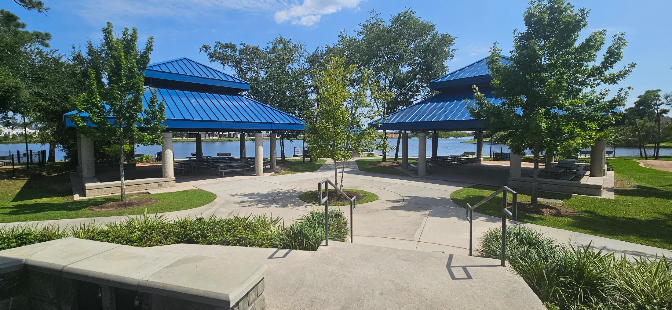 Two lakeside pavilions with cobalt-blue standing-seam roofs facing each other across a paved plaza, picnic tables beneath each, open water behind