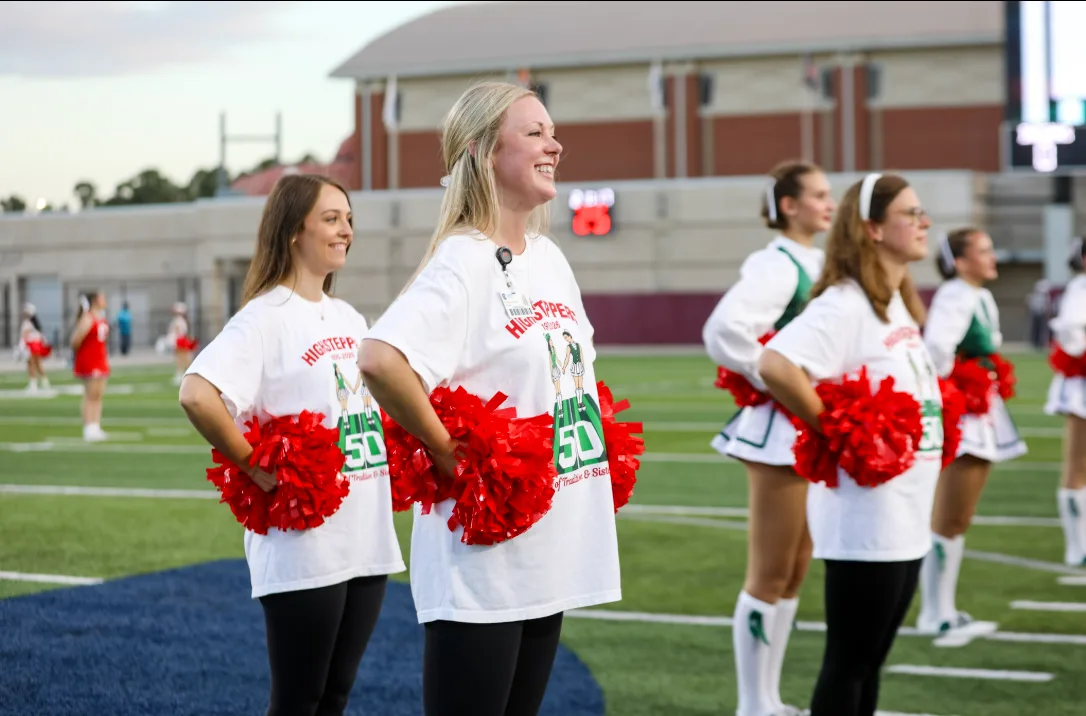 Alumni dancers in Highsteppers 50th anniversary shirts stand on the field at Woodforest Bank Stadium holding red pom-poms