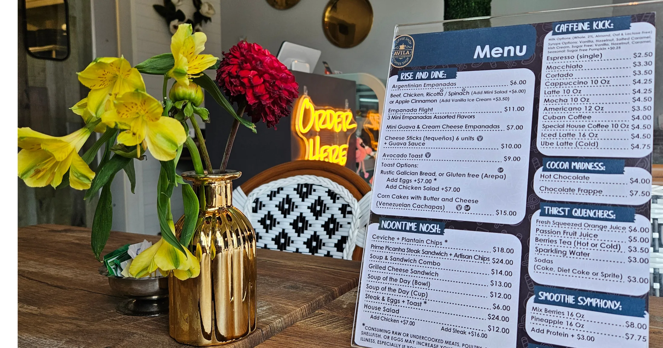 Table at Avila Coffee Shop with gold vase of yellow and red flowers, menu card, and neon Order Here sign glowing in the background