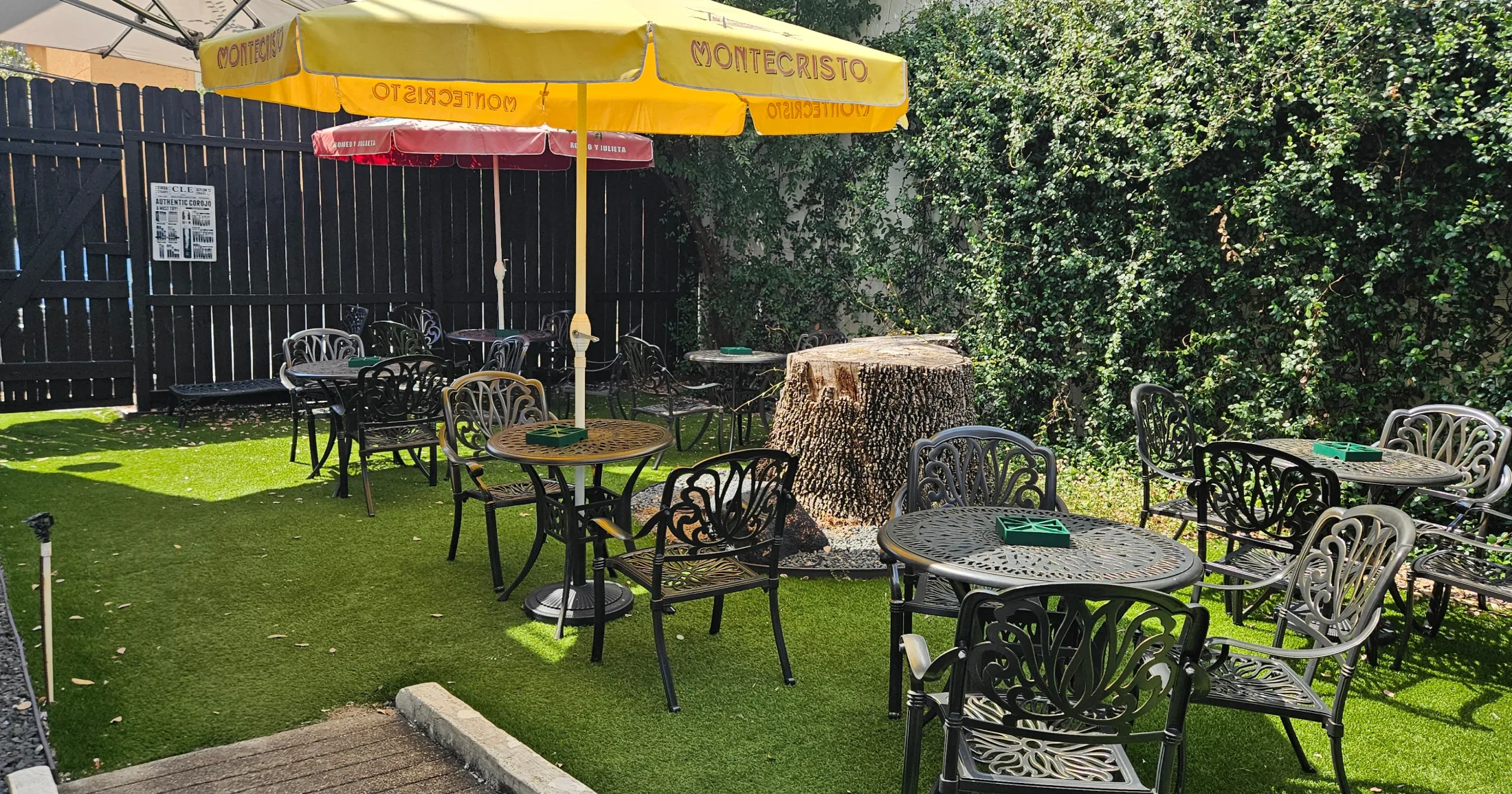 Outdoor cigar garden with cast-iron tables, Montecristo and Romeo y Julieta umbrellas, and a large tree stump centerpiece