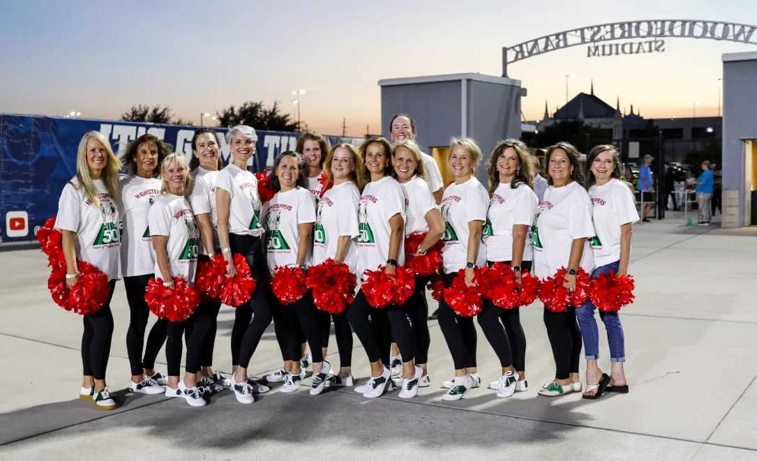 A large group of TWHS Highsteppers alumni in matching 50th anniversary shirts pose together holding a 2000s decade sign