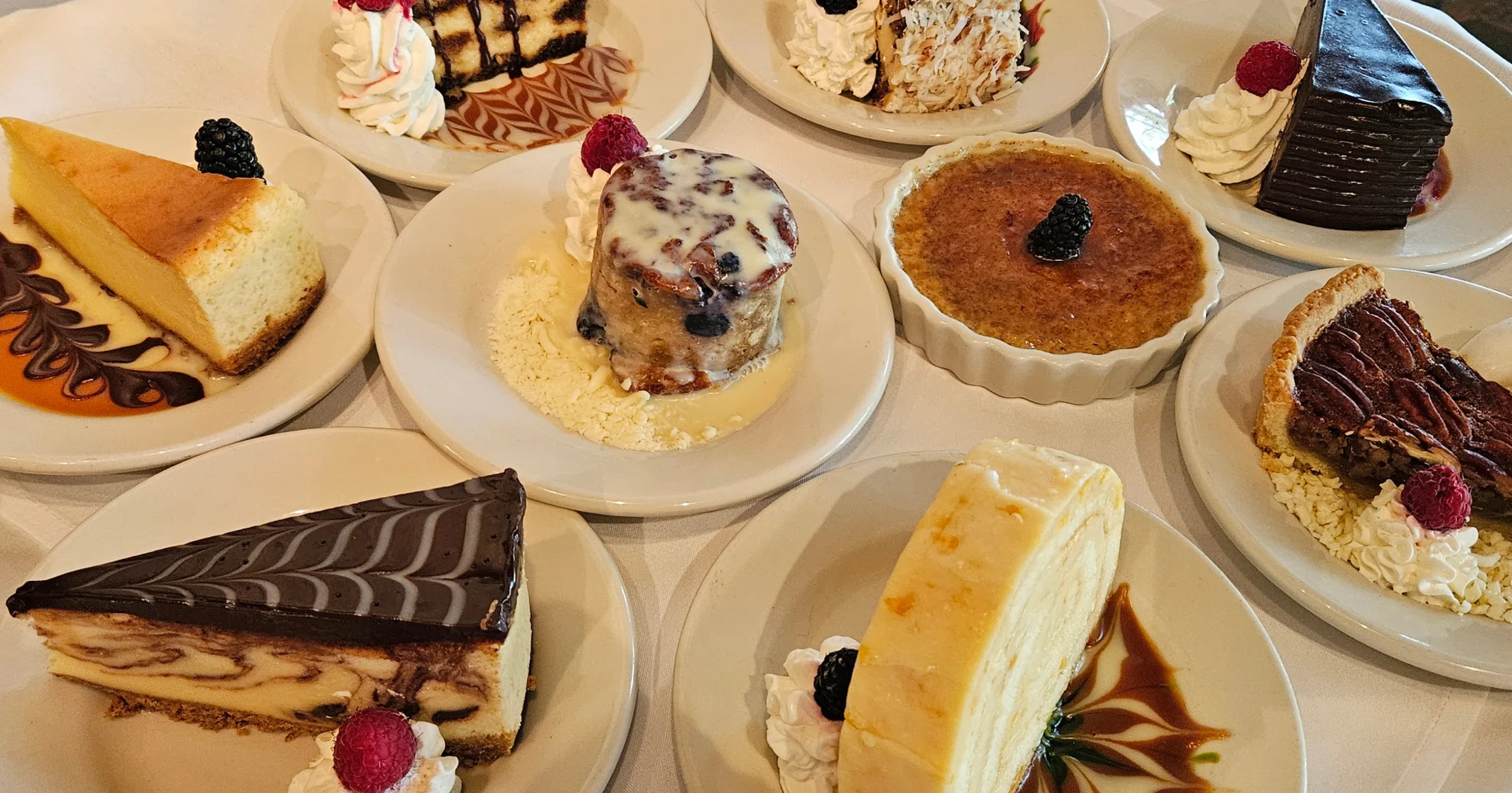 Nine dessert plates spread across a white tablecloth including cheesecakes, creme brulee, bread pudding, and pecan tart