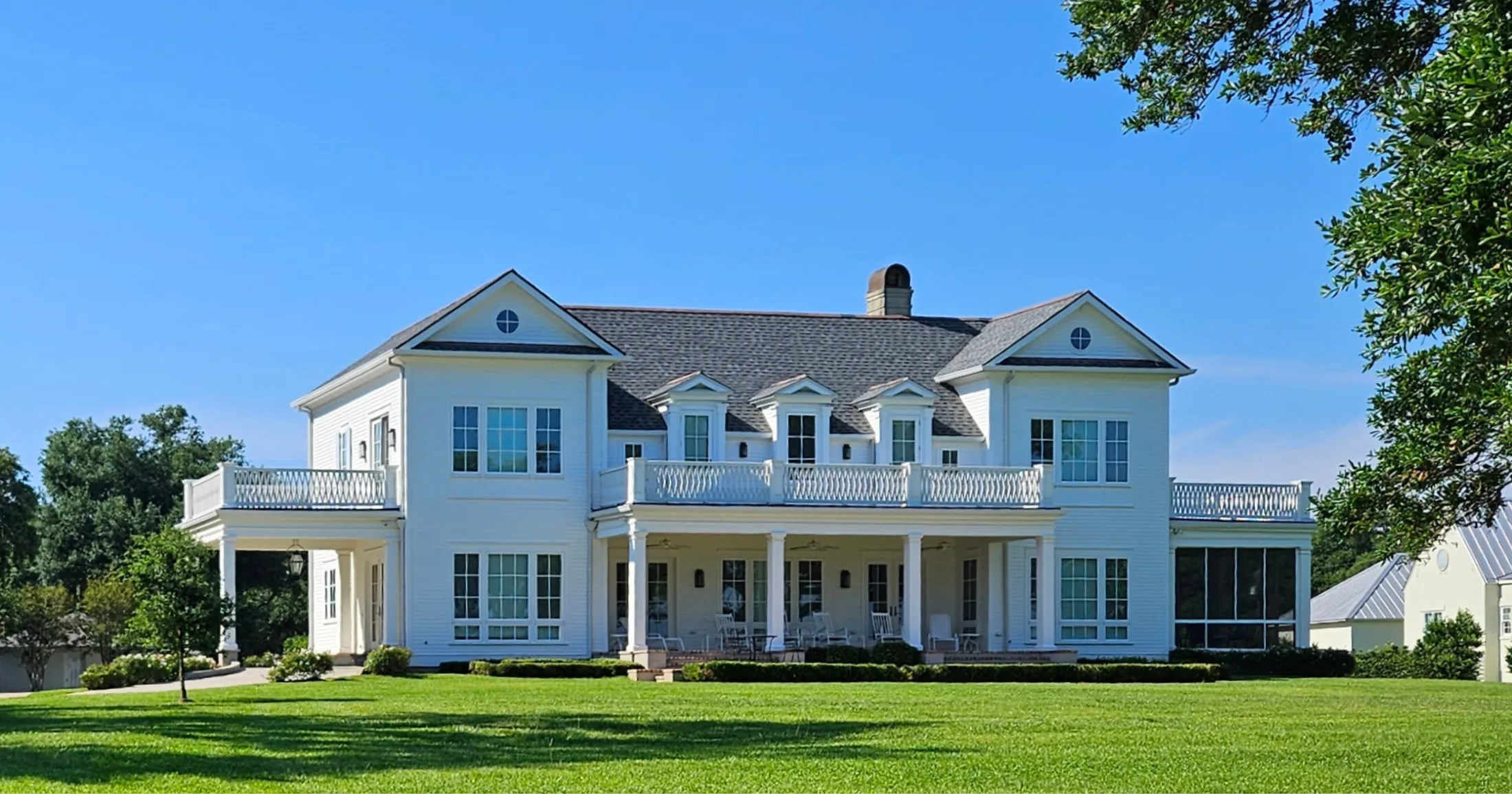 A grand white two-story Southern colonial home with full-width wrap-around porches, white balcony railings, and manicured grounds along the Gulfport beachfront.