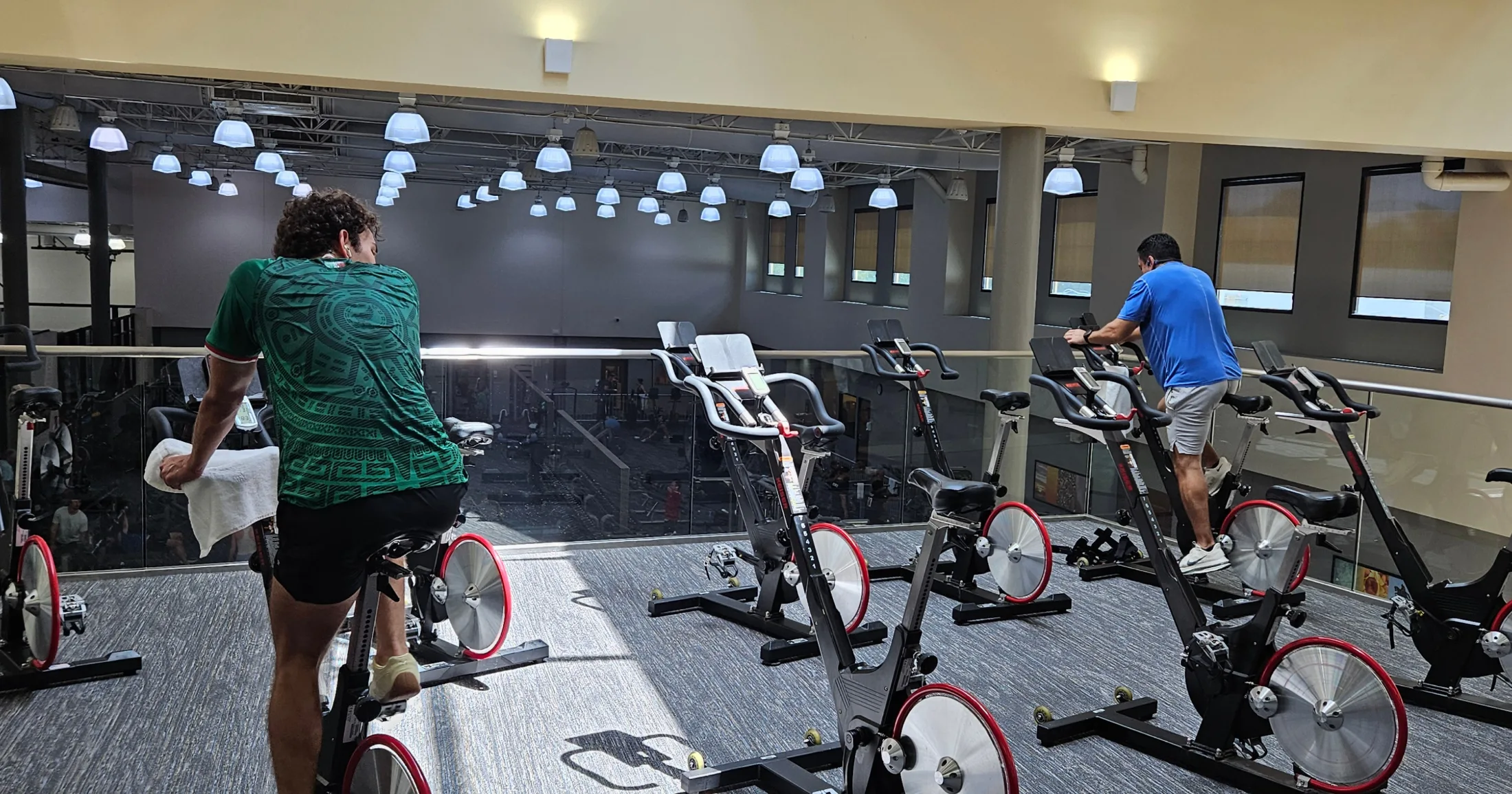 Two members on cycle bikes on the Villa Sport mezzanine level, overlooking the weight floor below