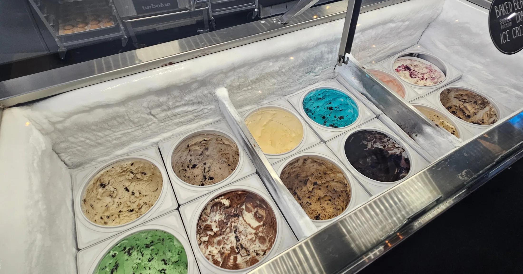 Super premium ice cream case at The Baked Bear showing more than a dozen flavors including green mint, blue Cookie Monster, dark chocolate, and berry swirl