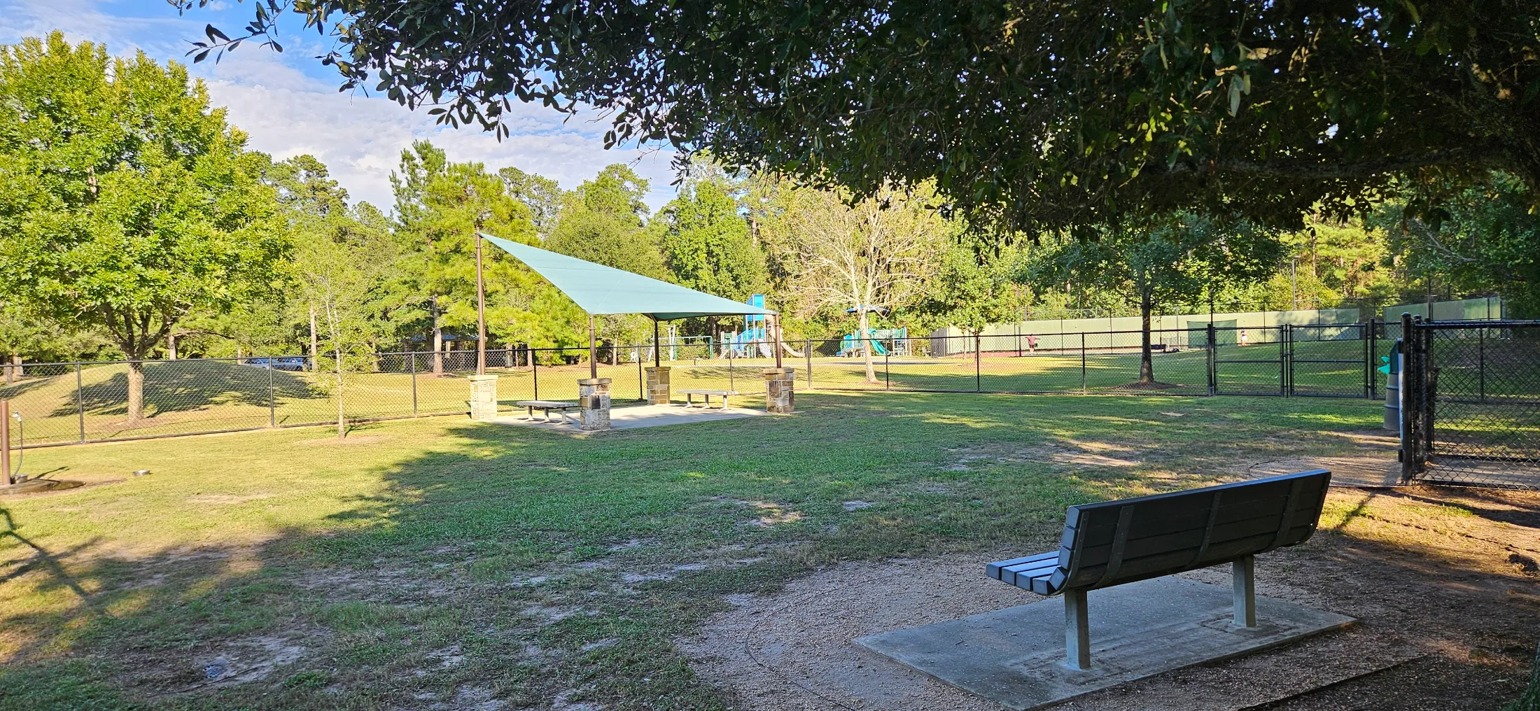 Cattail Park dog park bench in the foreground, green shade sail and stone pillar shelter visible in the middle distance, playground and tennis courts seen through the fence line beyond