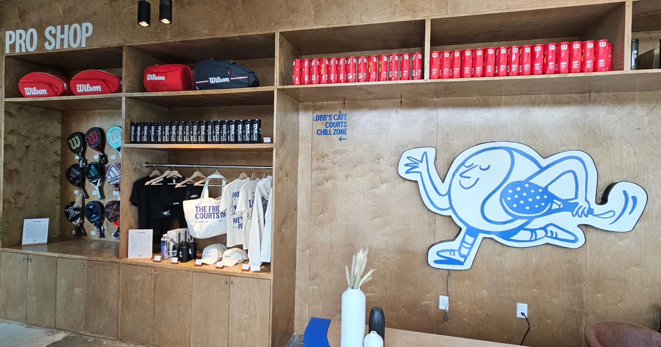Lobb's Padel pro shop with Wilson rackets and bags on plywood shelving, branded apparel on a rack including tees, totes, and caps, canned drinks on the top shelf, and the Lobb's mascot graphic on the wall beside a directional sign for Lobb's Café, Courts, and Chill Zone
