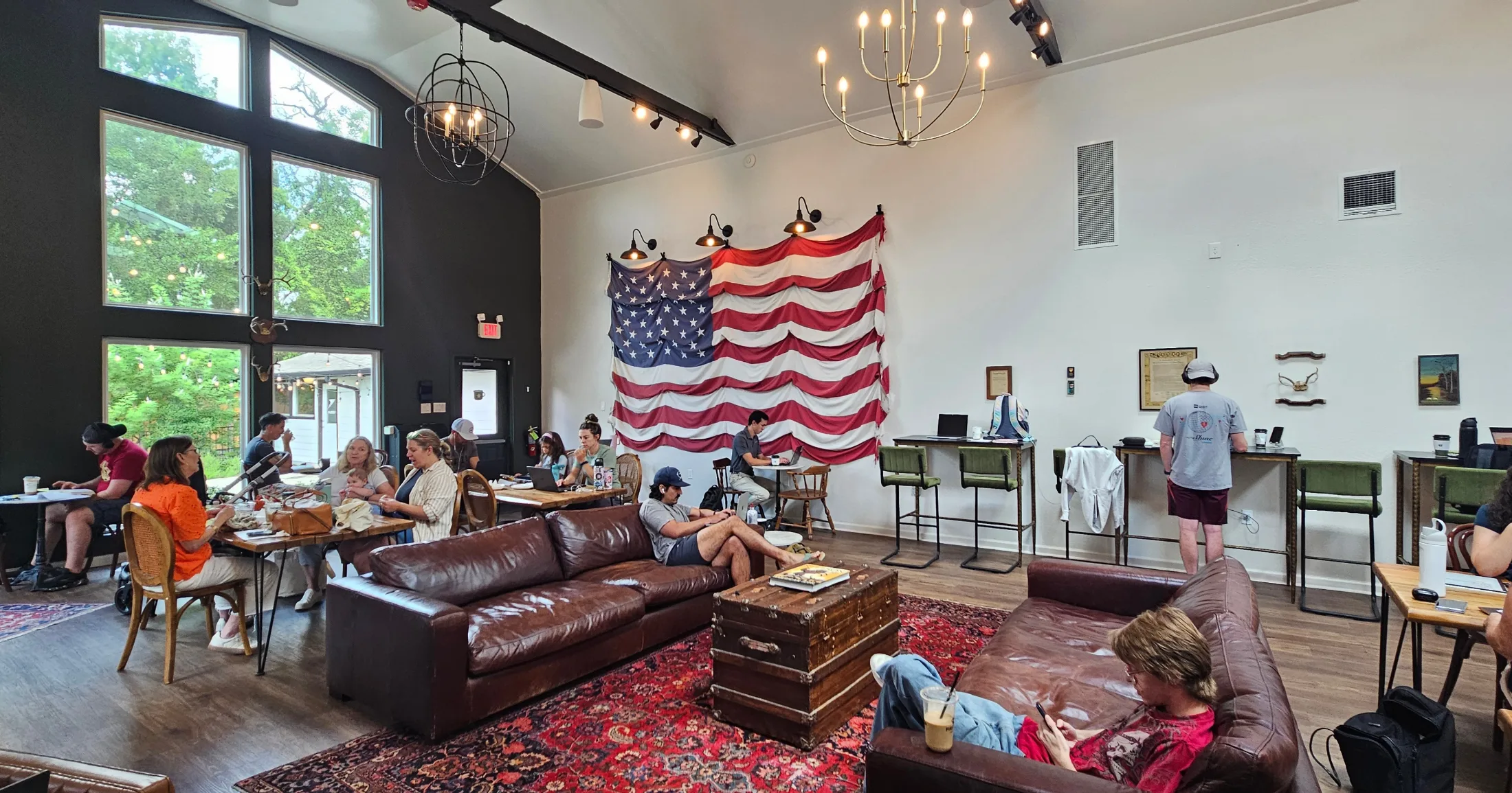 Interior of First Cup Coffee Co. showing leather sofas, dining tables full of guests, an American flag on the wall, and tall windows overlooking trees