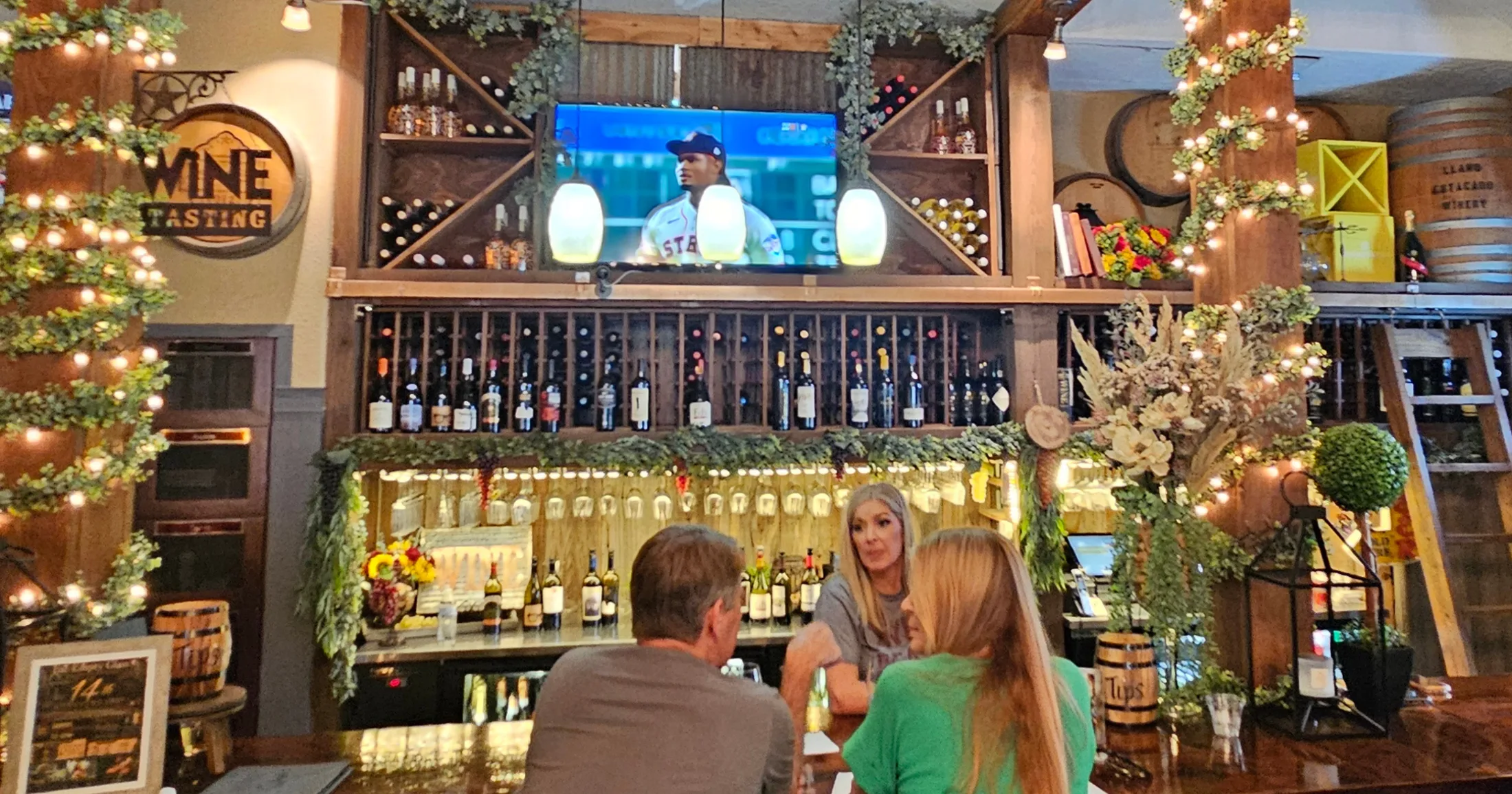 Full bar scene at The Empty Glass with guests seated at the bar, Astros game on the overhead TV, wine racks and barrel decor throughout