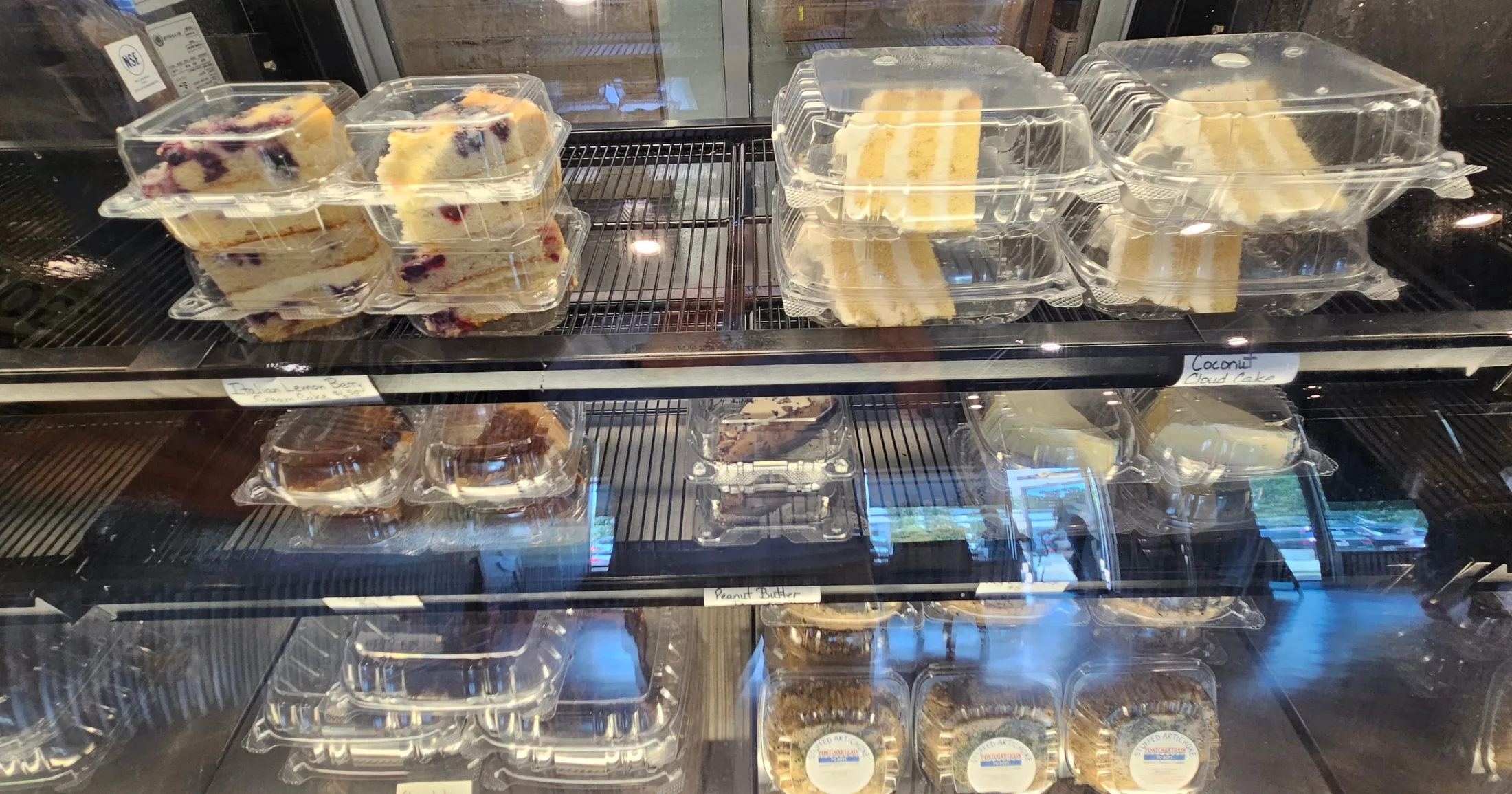 Dessert case displaying slices of Italian lemon berry choux cake, coconut cloud cake, peanut butter cake, and other pastries